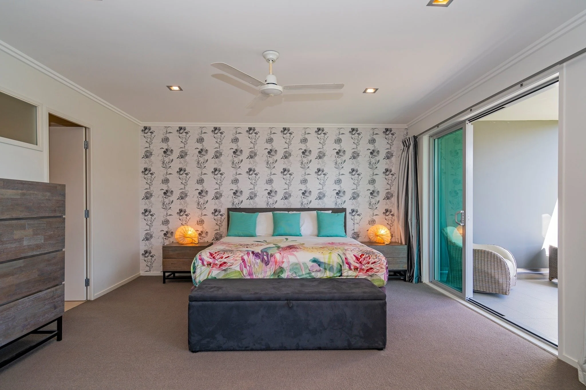 Bedroom with floral accent wall, wooden nightstands, turquoise pillows, and sliding glass door leading to balcony with outdoor furniture.