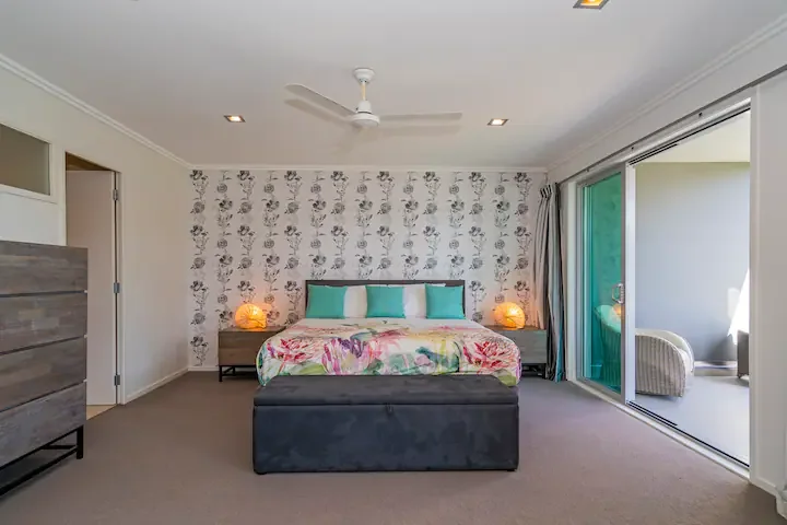 A bedroom with a bed decorated with blue pillows and a multicolored blanket, two nightstands with glowing lamps, floral wallpaper on the wall behind the bed, a ceiling fan, and sliding glass doors leading to an outdoor sitting area.
