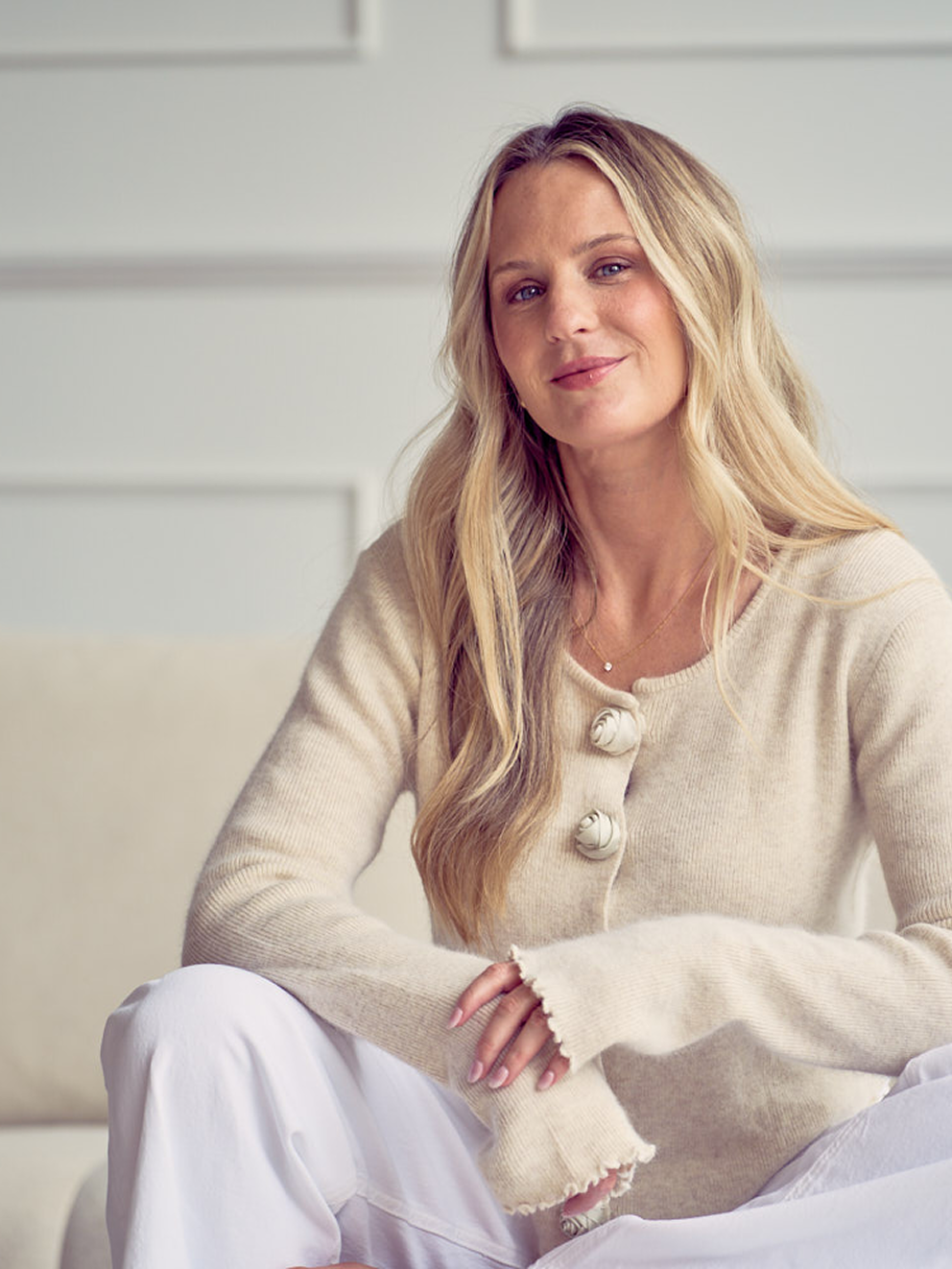 A woman with long blonde hair sitting on a sofa in a light-colored room, wearing a beige sweater and white pants, looking confidently at the camera.
