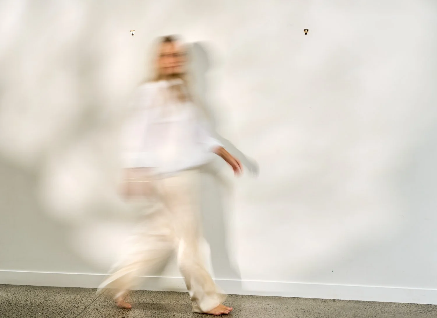 A person in white robes is walking or dancing blurred against a plain white wall, creating a motion effect.