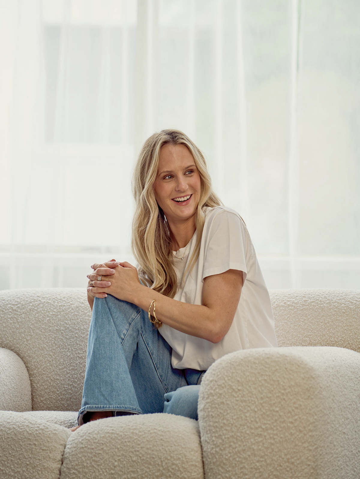 A woman with long blonde hair sitting on a beige couch, smiling, holding her knee with both hands, in a bright room with large windows in the background.
