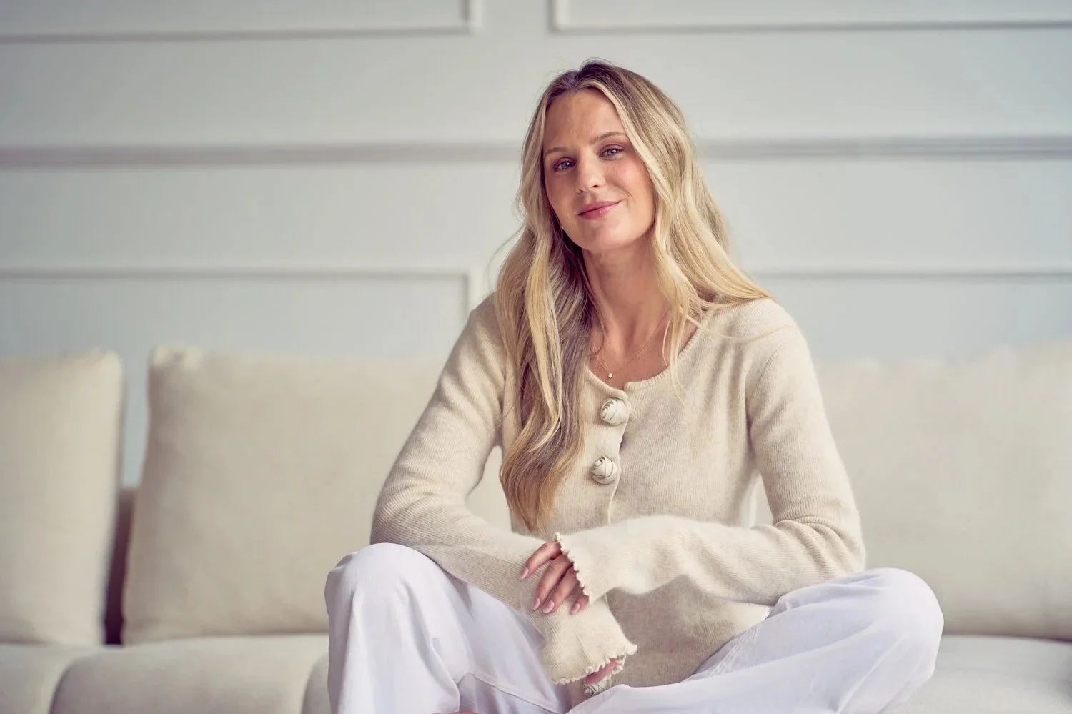 A woman with long blonde hair sitting cross-legged on a white sofa, wearing a beige sweater with large buttons and white pants, in a bright room with modern decor.