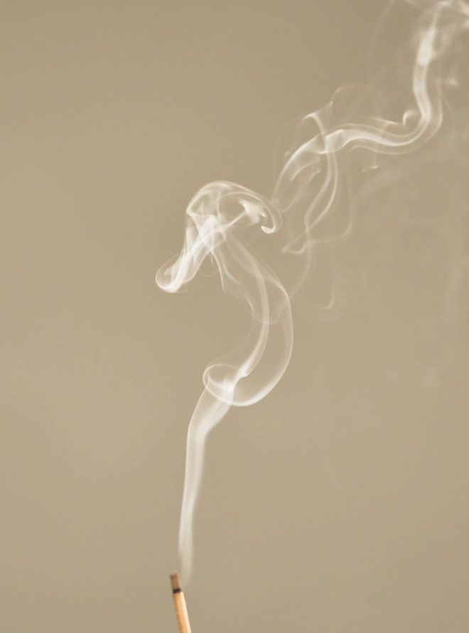 Thin trail of smoke rising from a lit incense stick against a plain, light brown background.