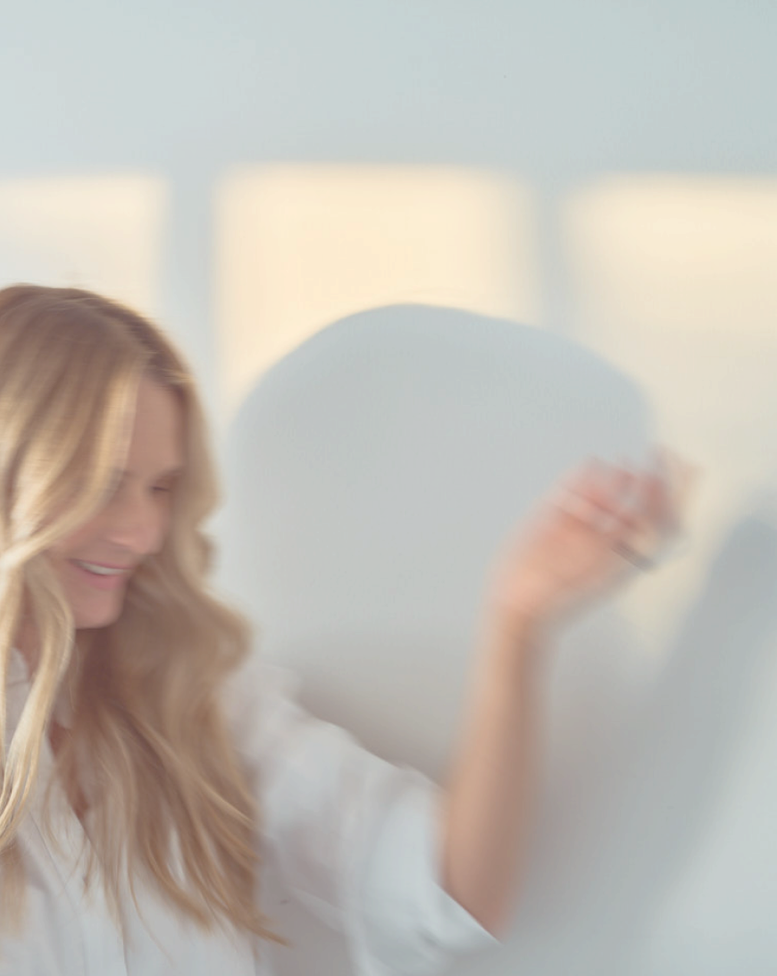 A woman with long blonde hair smiling and waving with her hand, blurred background with sunlight.