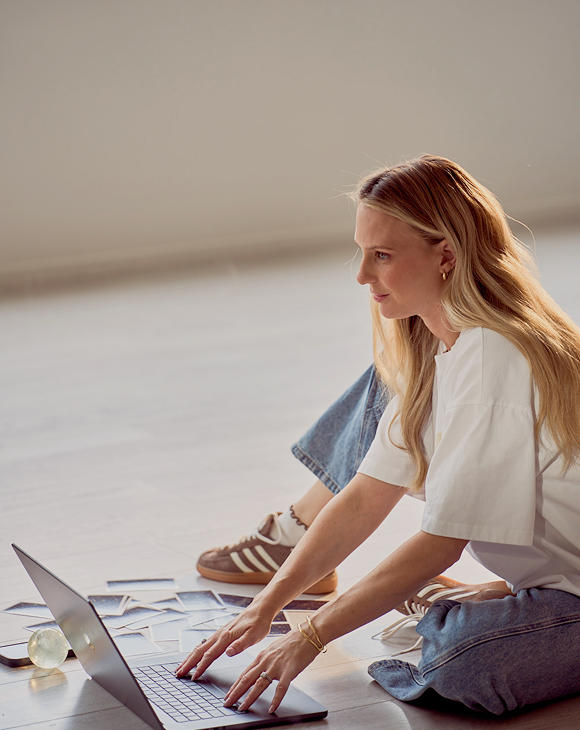 Woman with long blonde hair sitting cross-legged on a light-colored floor, using a laptop with photos spread around her.