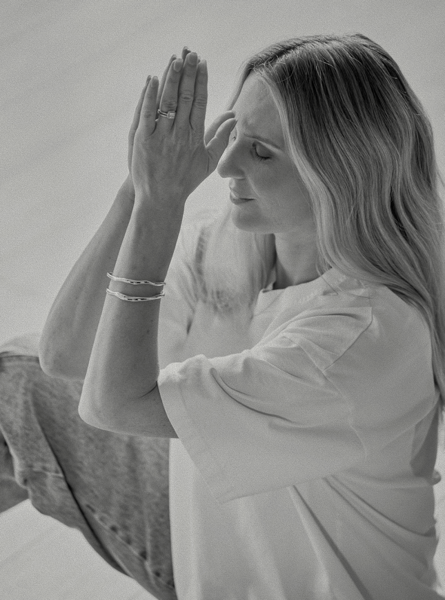 A woman with long hair and closed eyes holds her hands together near her face in a prayerful or meditative pose, wearing bracelets and a loose top.