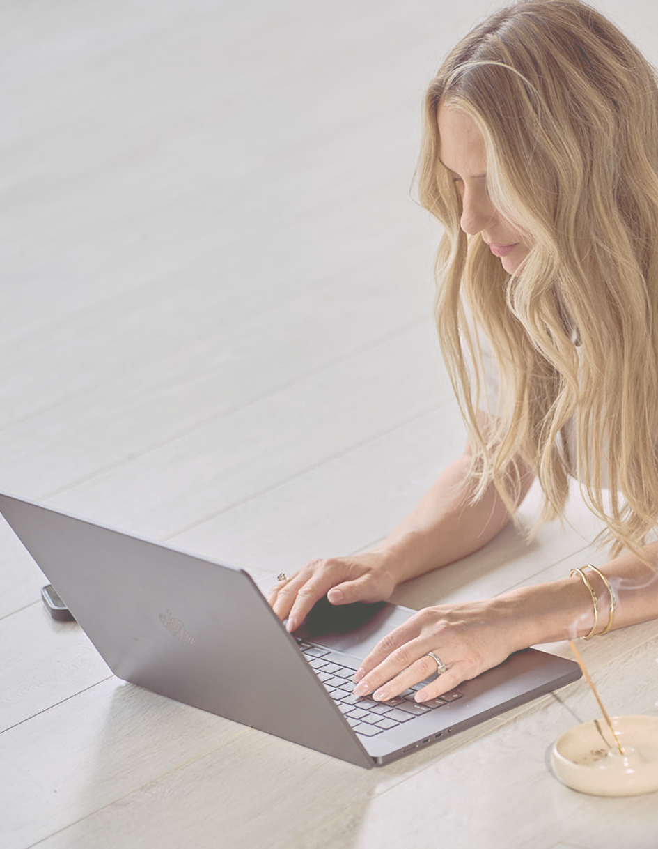 A woman with long blonde hair working on a silver laptop, sitting on a light wood floor.