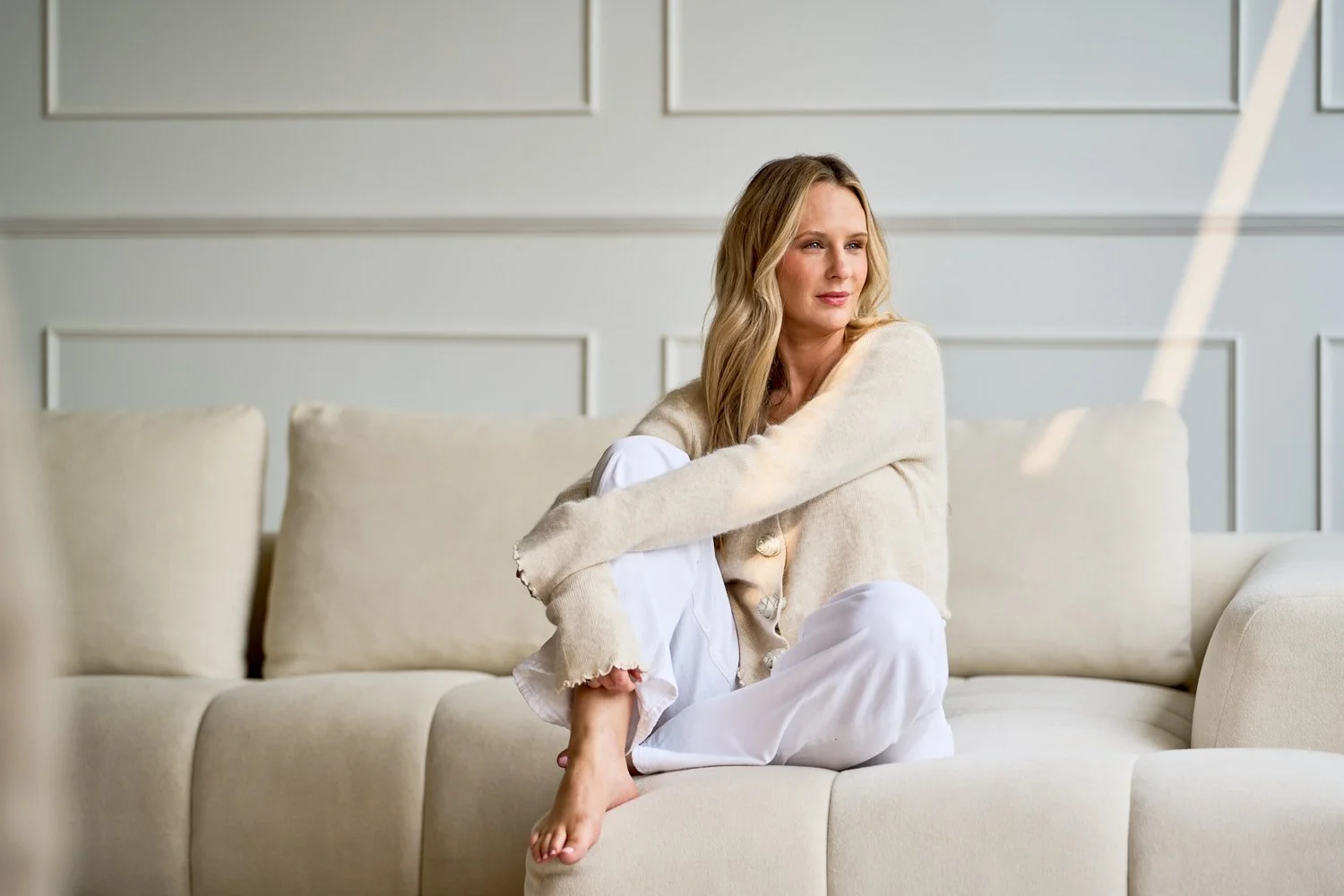 A woman with long blonde hair sitting on a beige sofa in a light-colored room, wearing a cream-colored cardigan and white pants, looking off to the side.