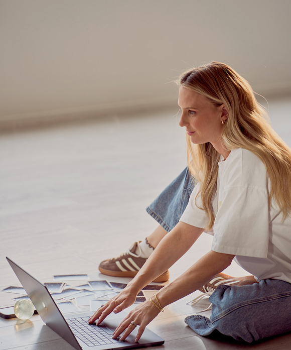 Young woman sitting on the floor using a laptop, with scattered papers around her, in a bright indoor space.