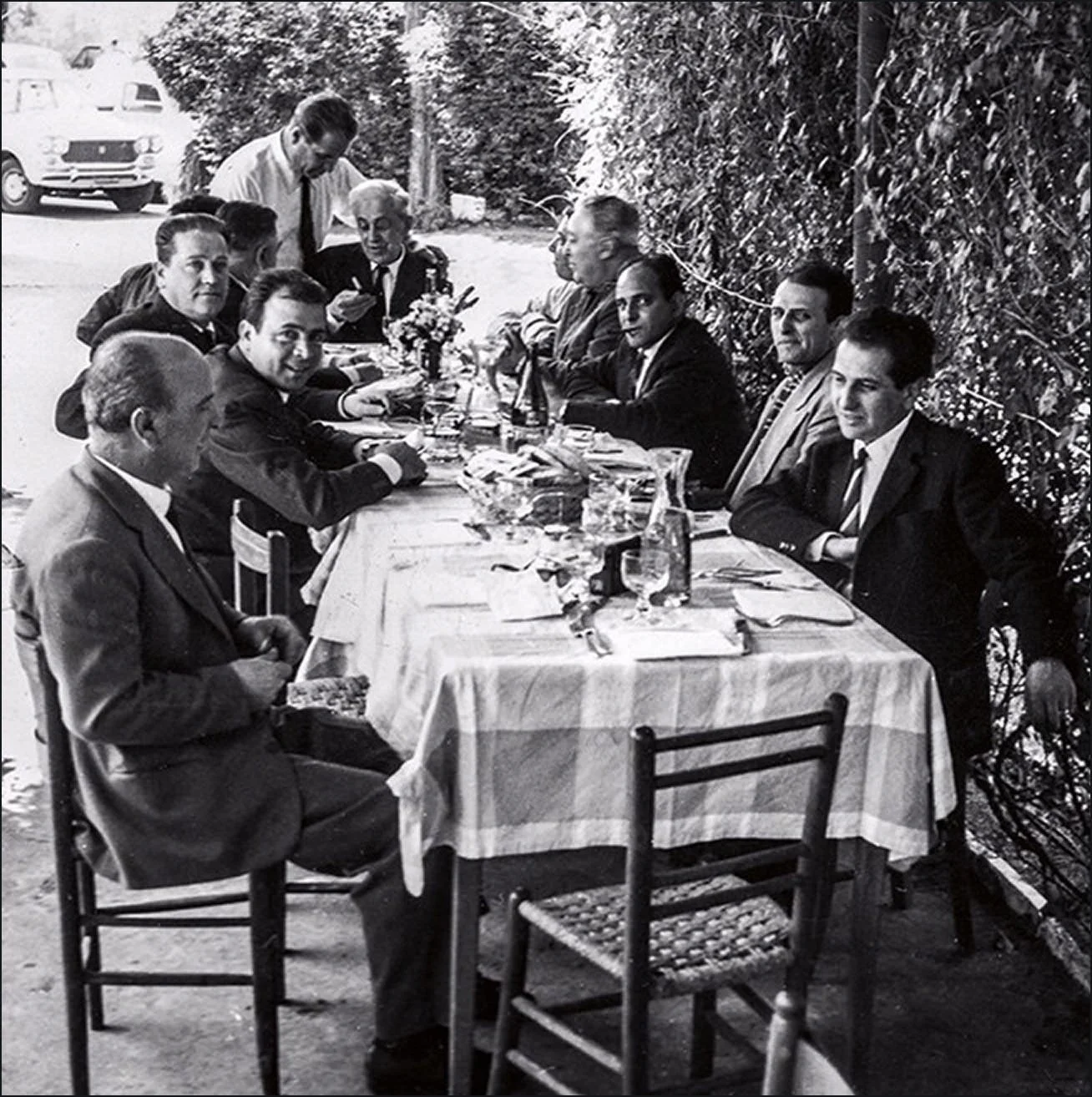 Black and white photo of a group of men in suits sitting at an outdoor dining table, with glasses, plates, and a floral centerpiece, against a hedge and parked cars in the background.
