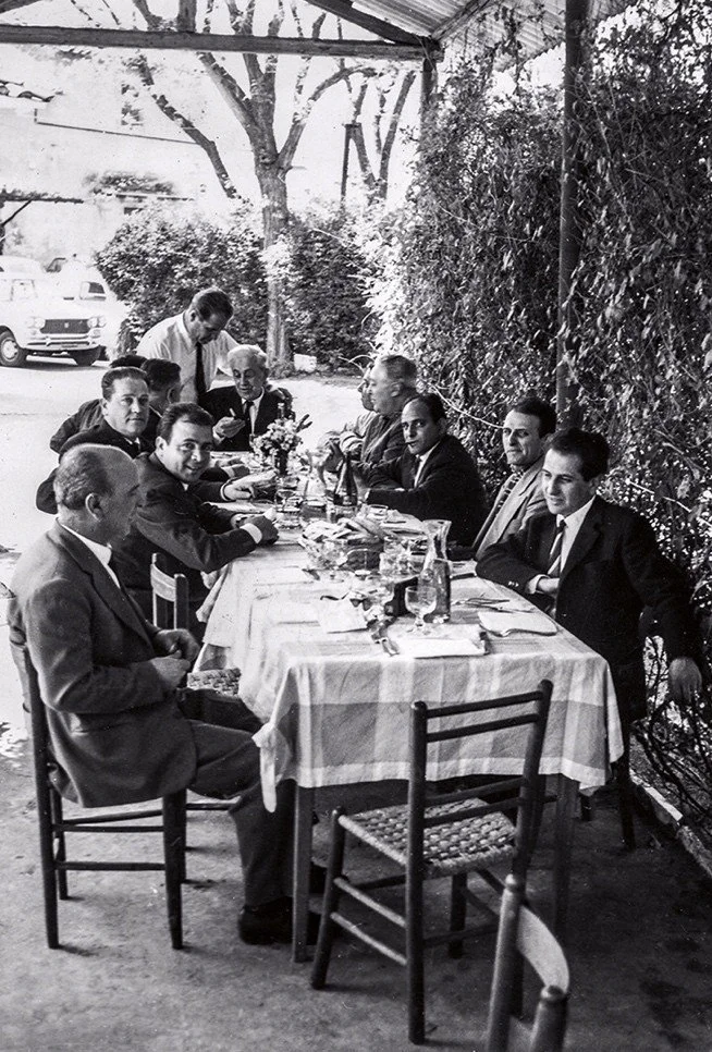 Black and white photo of ten men dressed in suits seated around a long table outdoors, with a server standing behind them. The table has a checkered tablecloth, drinks, and a small flower arrangement. There are trees and bushes in the background.