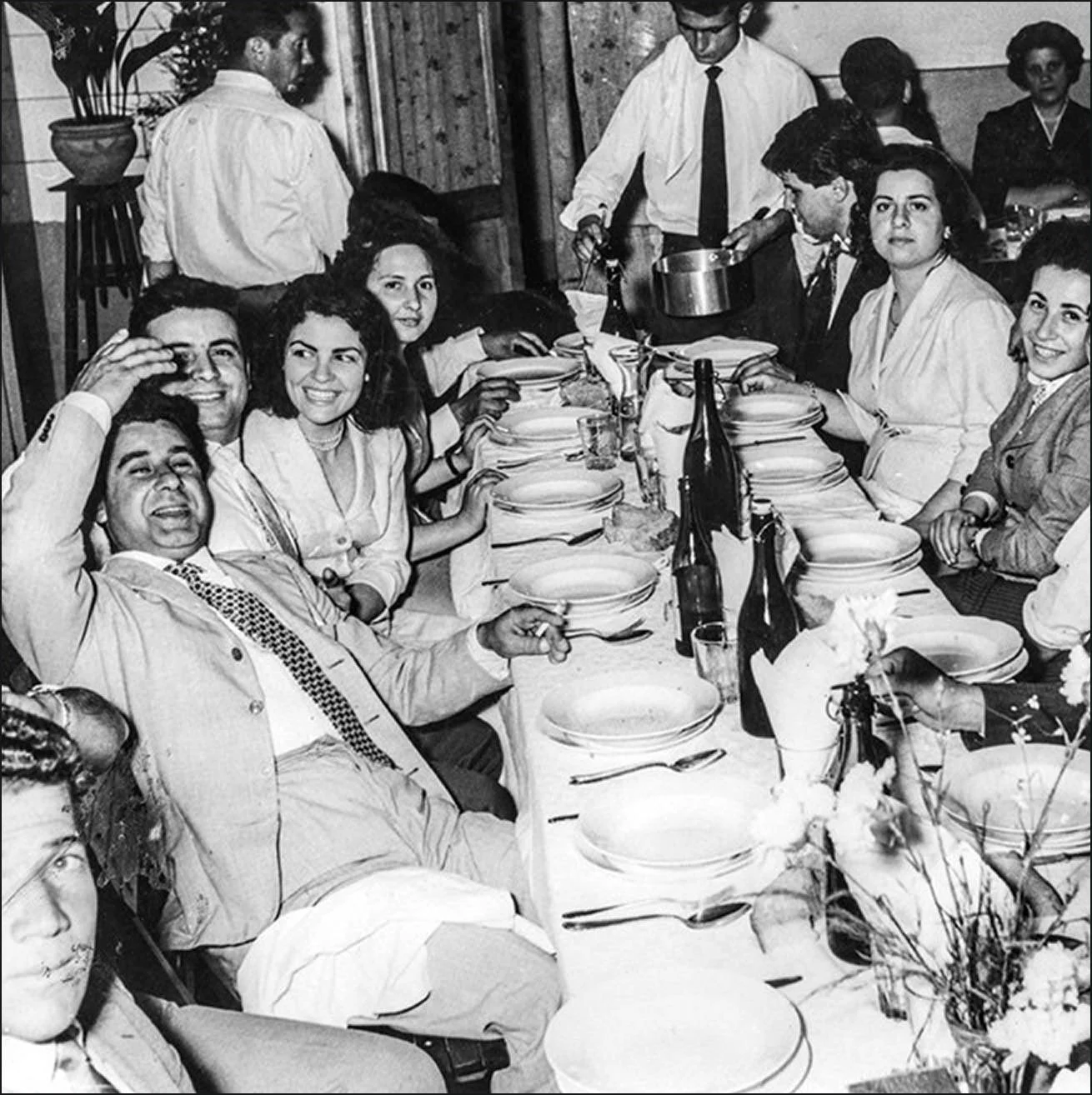 Black and white photo of a group of people sitting at a dinner table, smiling and enjoying a meal together, with some men in suits and women in dresses.