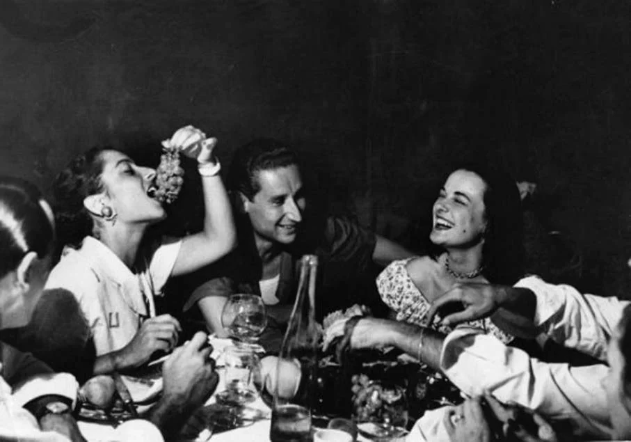 Group of people enjoying a meal together in black and white, with one woman lifting a glass and smiling, others engaged in conversation and laughter.