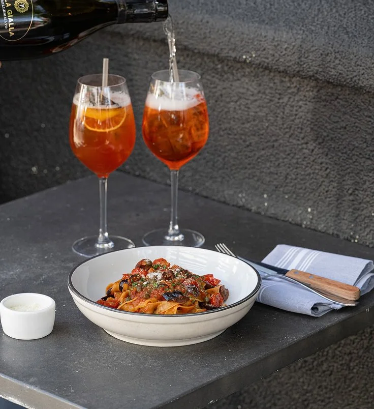 Two glasses of iced tea or cocktail with orange slices being poured, a bowl of pasta with tomato and vegetable sauce, a small dish of white sauce, and a fork with a knife on a napkin on a gray outdoor table against a dark stone wall.
