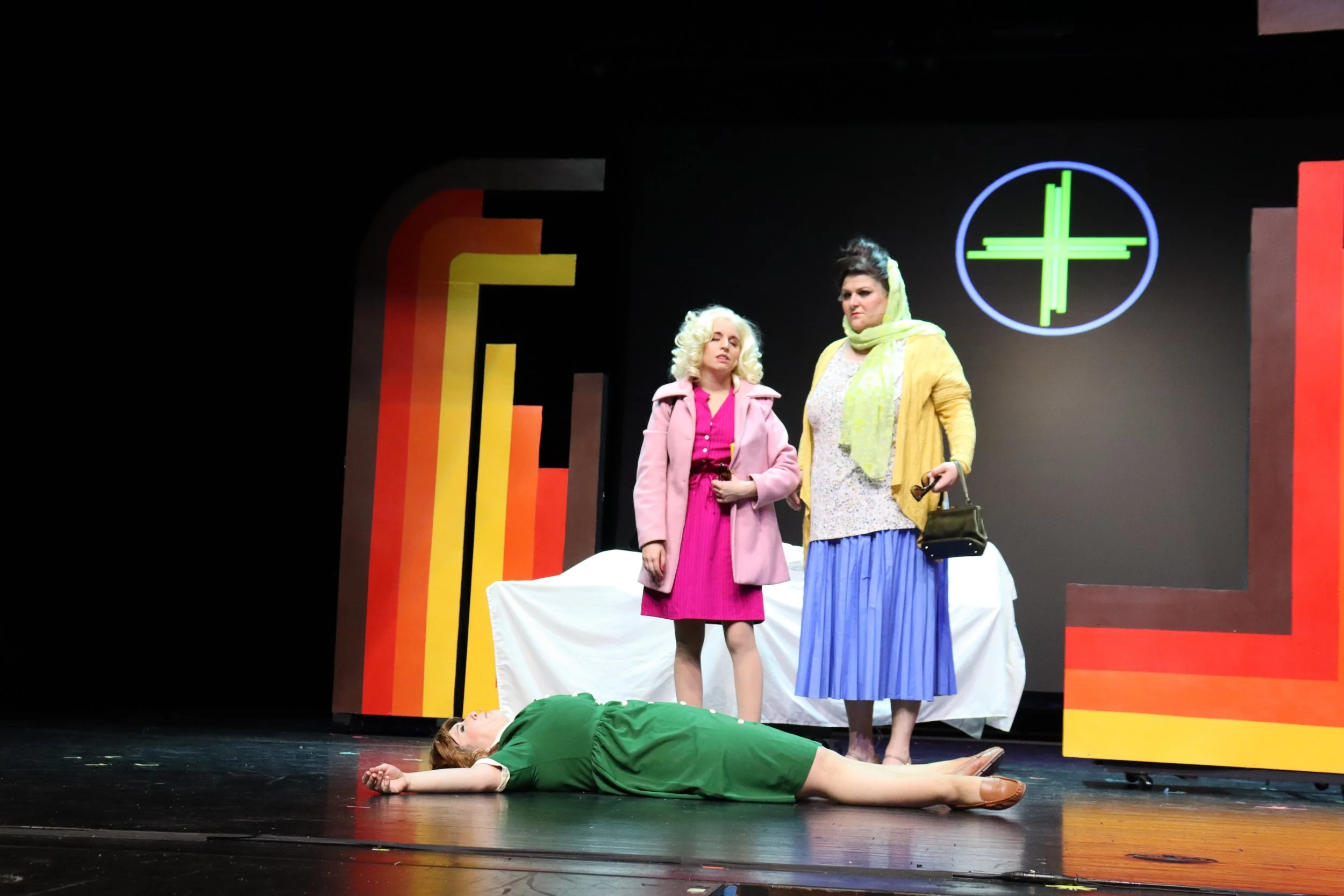 Three women on stage in a theatrical play, with one lying on the floor and two standing, against a backdrop with neon symbols and colorful geometric panels.