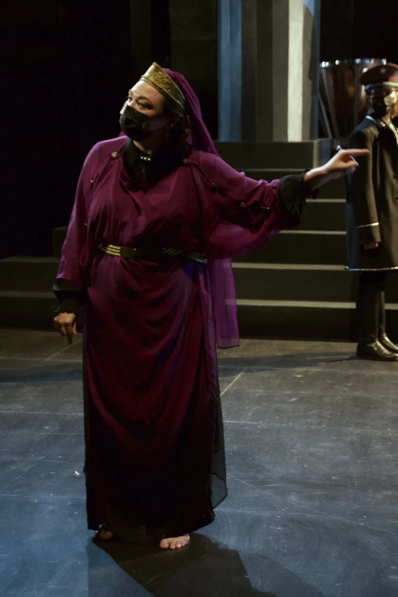 Woman dressed in a purple gown and a headpiece, wearing a black face mask, standing on stage and pointing to the right.