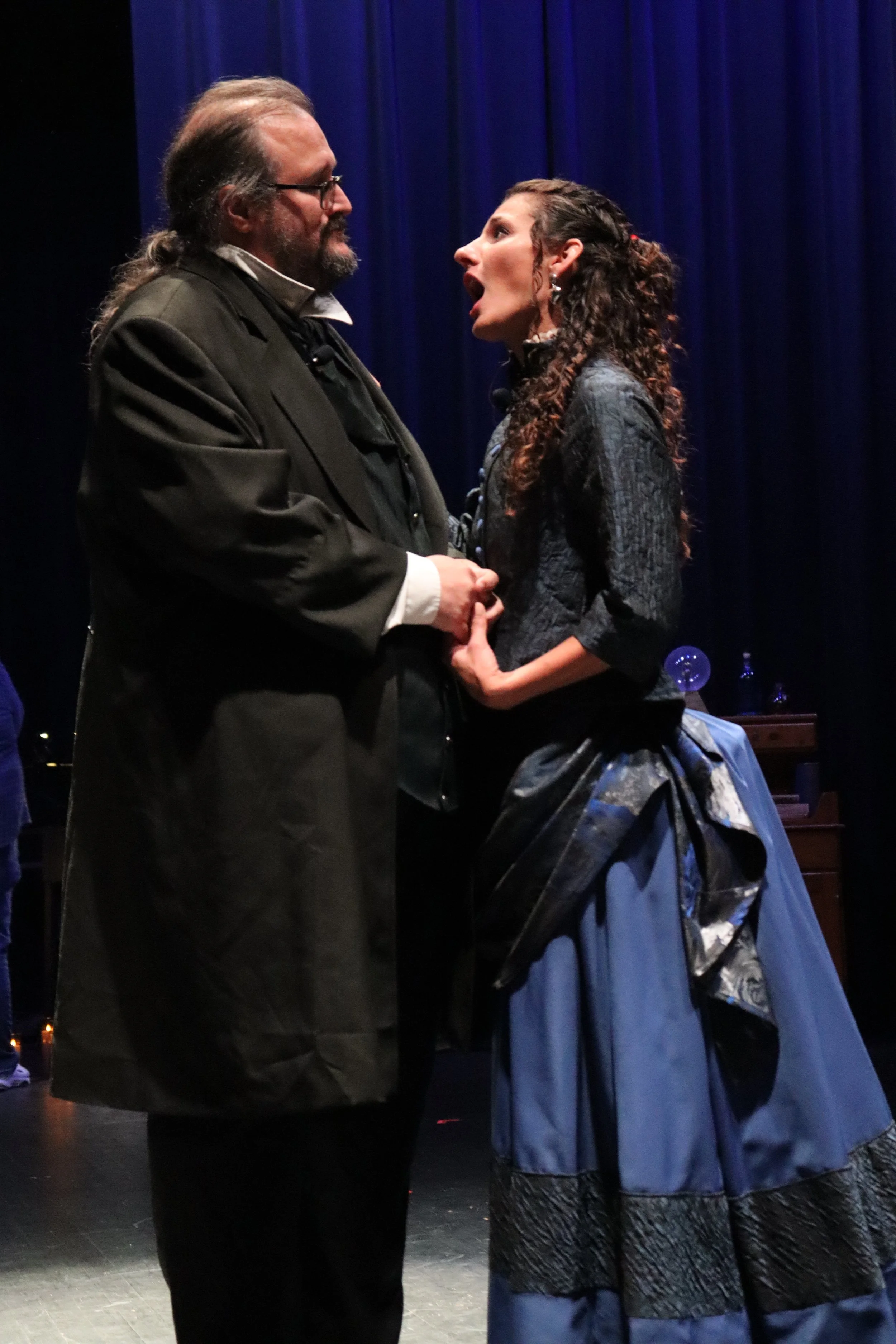 Two actors on stage, a man dressed in historical 19th-century attire, and a woman in a Victorian-era dress, holding hands and looking at each other, with blue curtains in the background.