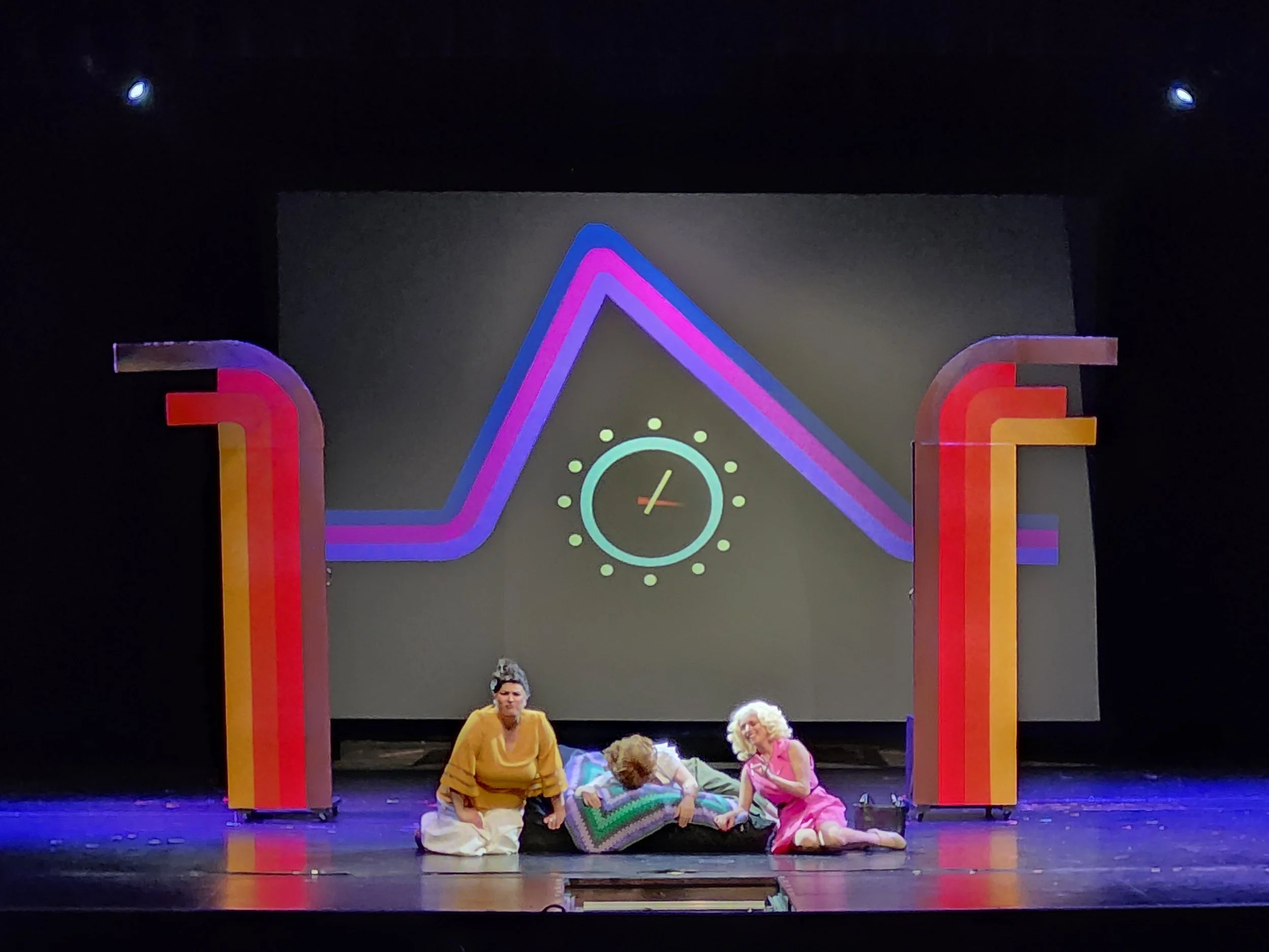 Three performers on stage during a theatrical play, with two women kneeling and one lying on the floor, holding a colorful quilt. A large clock icon with a pulse line is on the screen behind them, framed by brightly colored, curved structural element