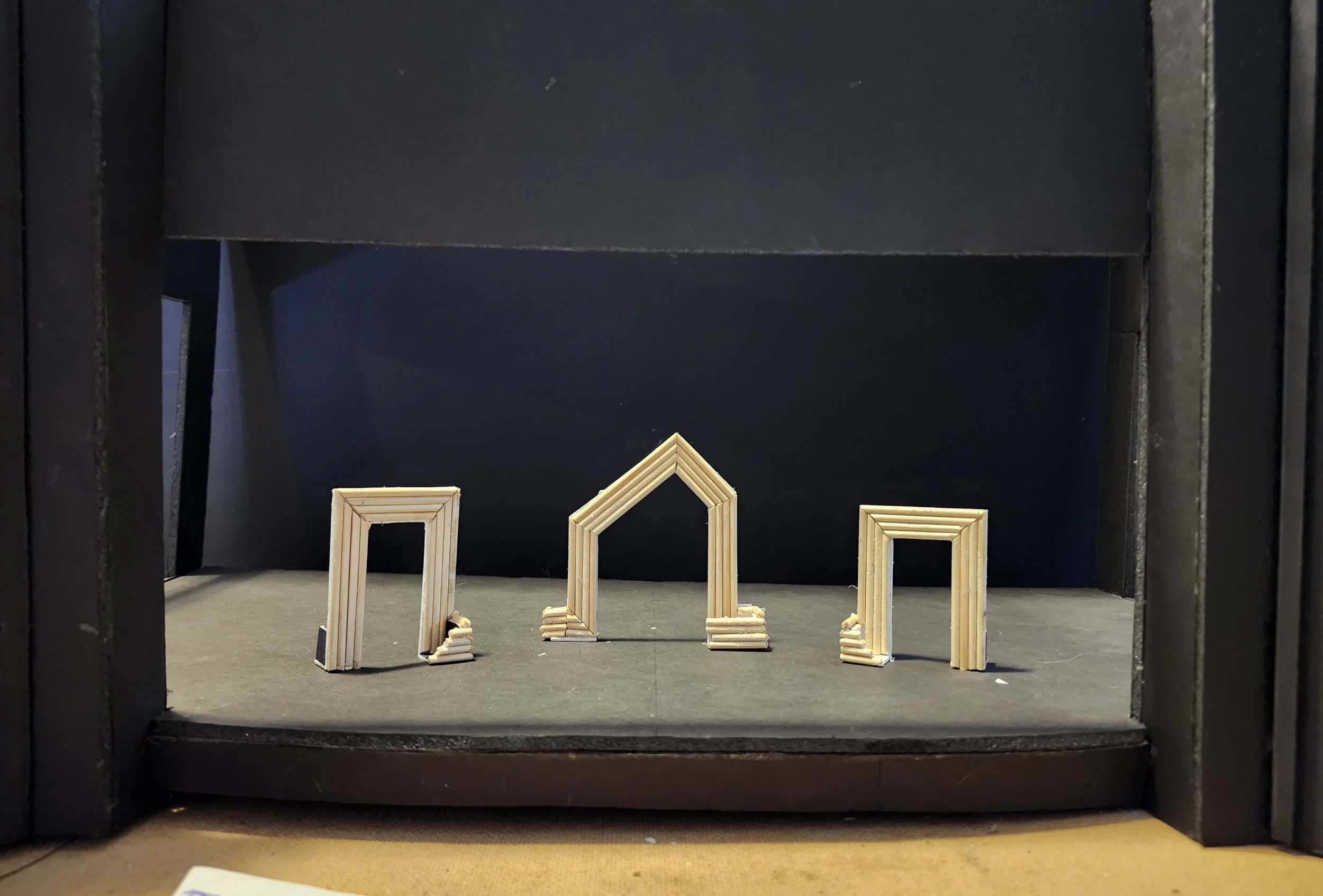 Miniature wooden arch and doorway models displayed on a dark stage within a black frame.