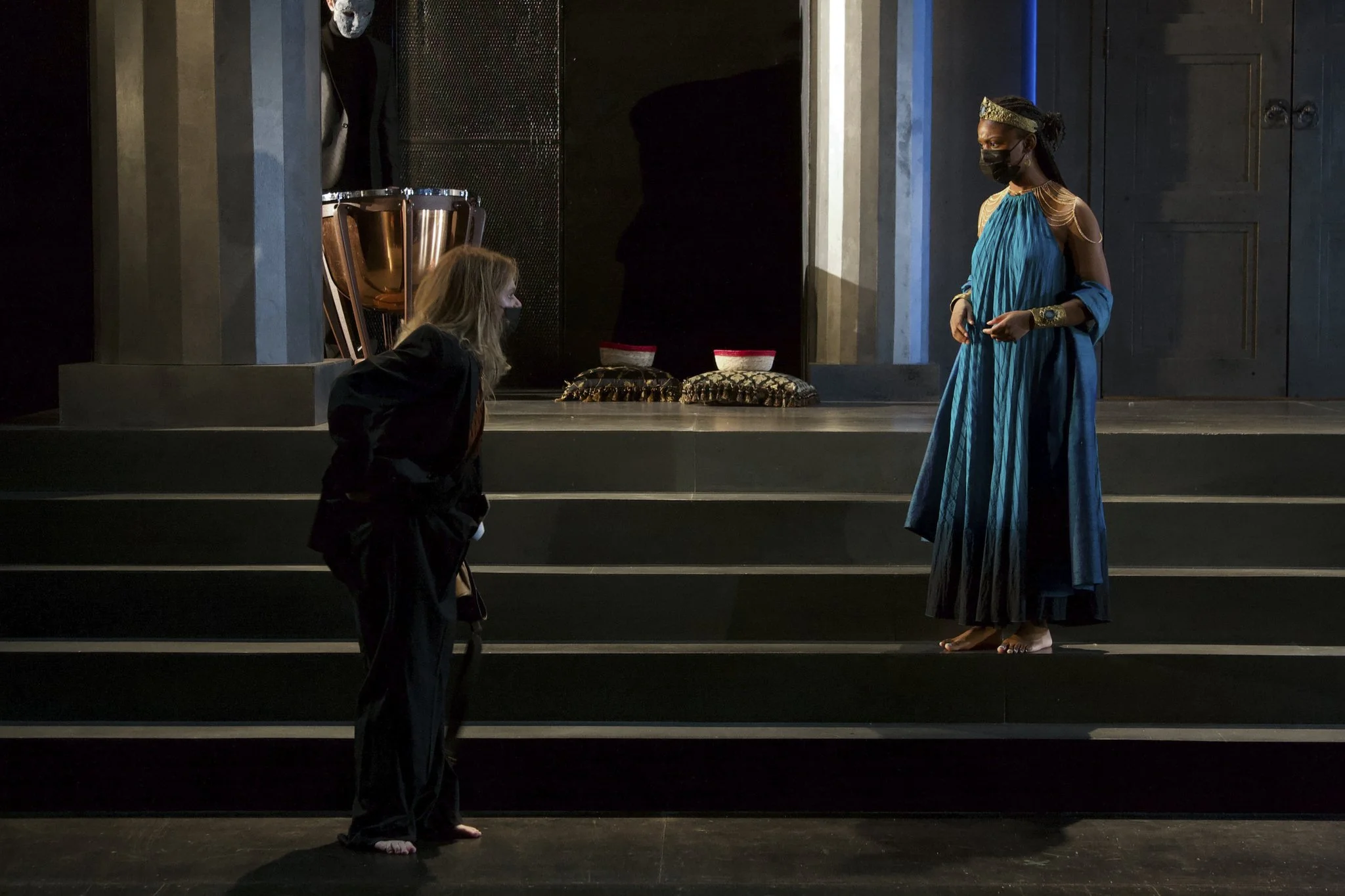 Two women on stage, one standing in a bright blue dress with arm jewelry and the other kneeling in a black outfit, engaged in a conversation. A person with a painted white face sits in the background near musical percussion instruments.