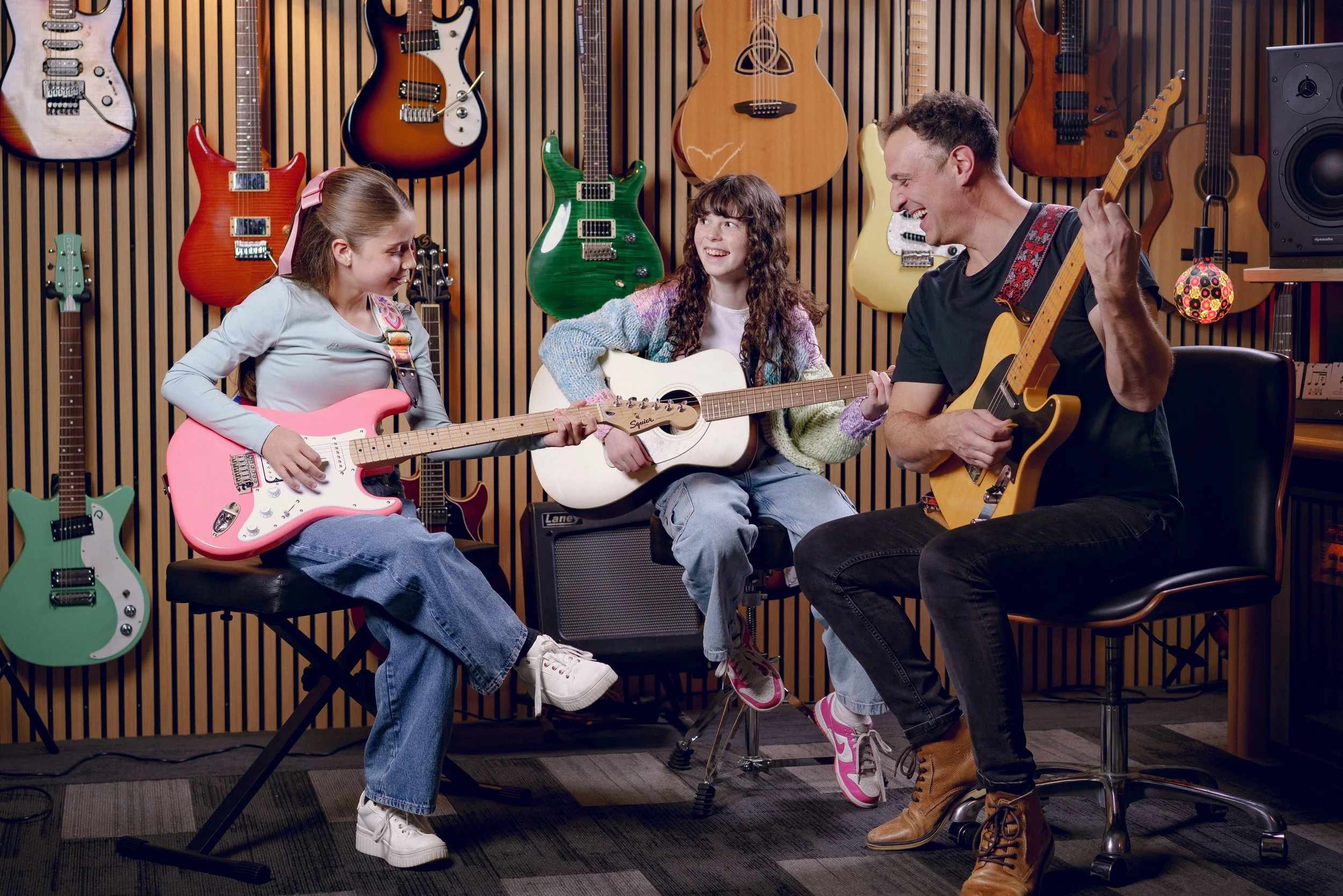 Three people, a man and two girls, are sitting with guitars in a music studio. They are smiling and playing their guitars, with a background of colorful guitars hanging on the wall.