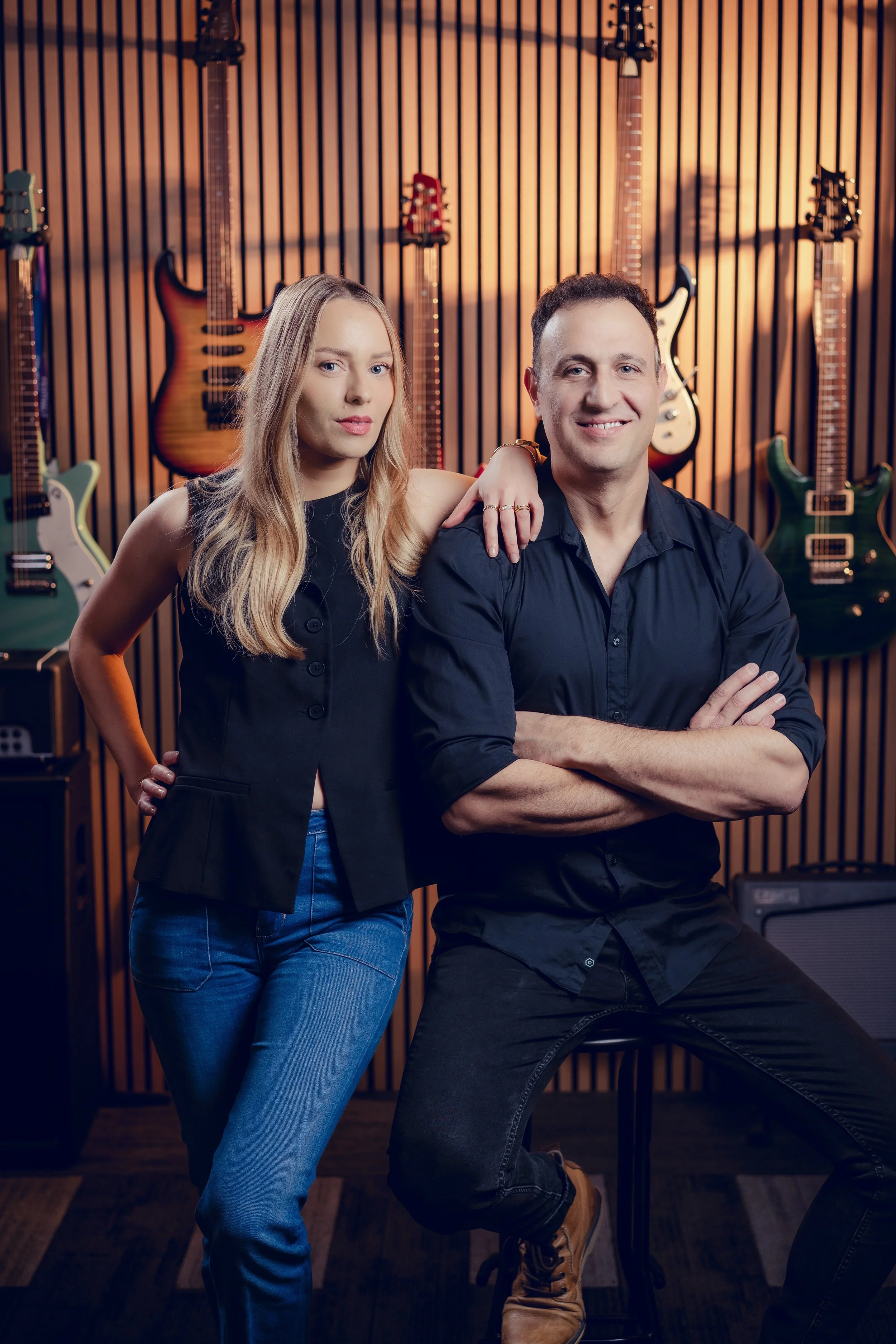A man and woman pose in a music studio with guitars hanging on the wall behind them.