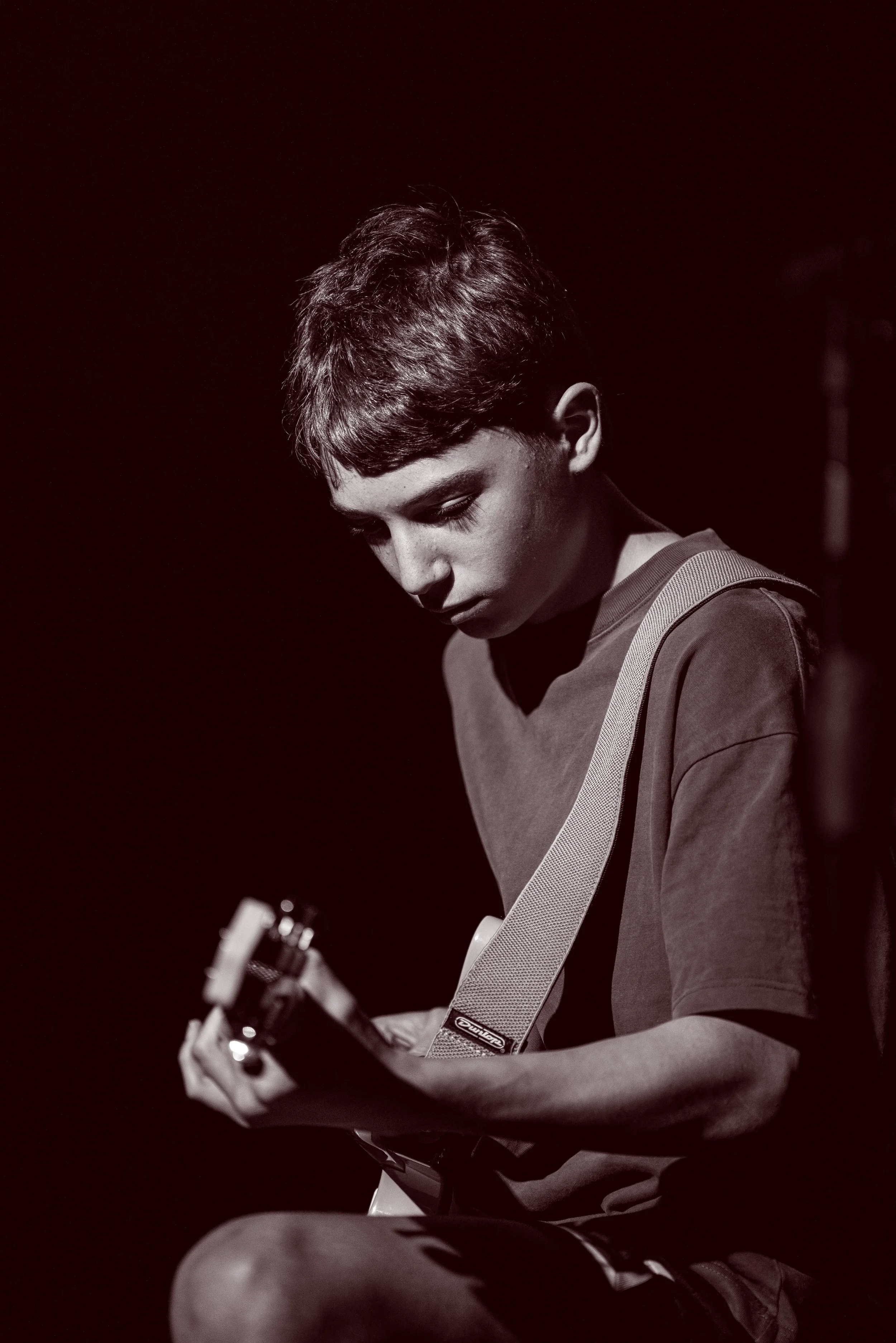 A young boy with short hair playing an acoustic guitar on stage, illuminated by a spotlight, with a dark background.