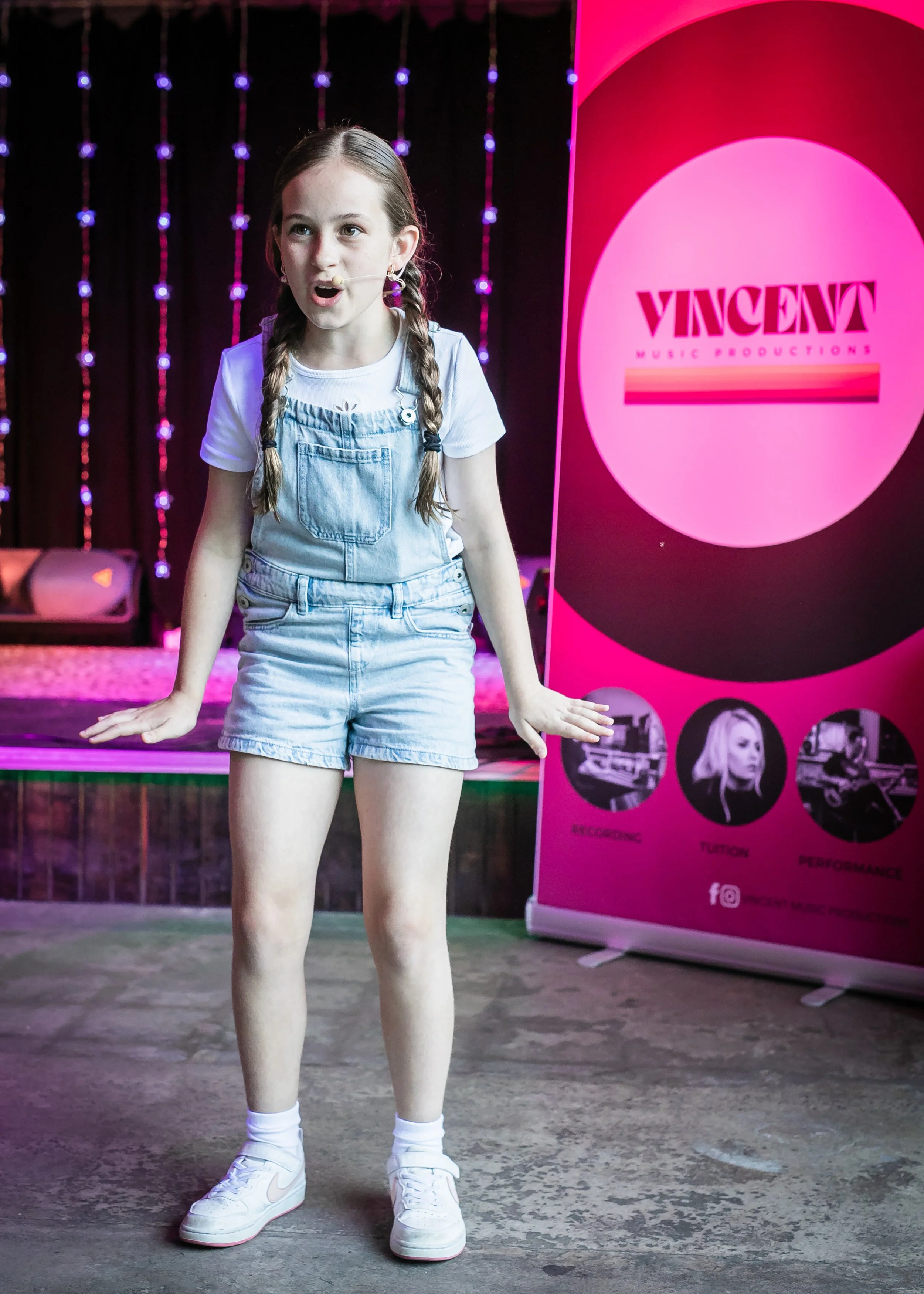 A young girl with braided hair standing on a stage, speaking or performing, with a pink and black banner behind her displaying 'VINCENT Music Productions' and icons for recording, vocals, and performance.