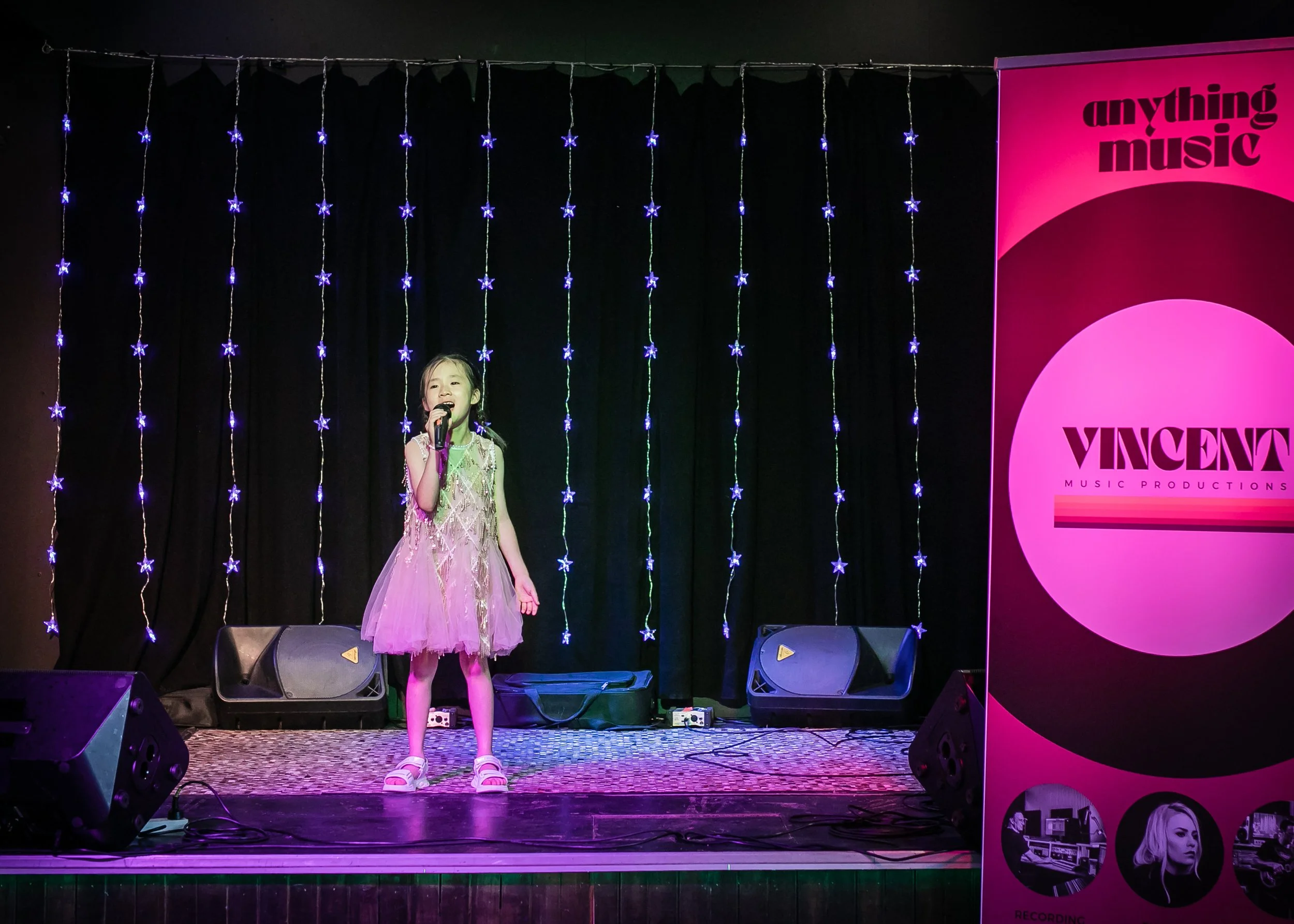 A young girl in a pink dress singing into a microphone on stage. Black curtains and blue star-shaped string lights are in the background. A large pink and purple banner on the right displays "VINCENT" and "anything music".