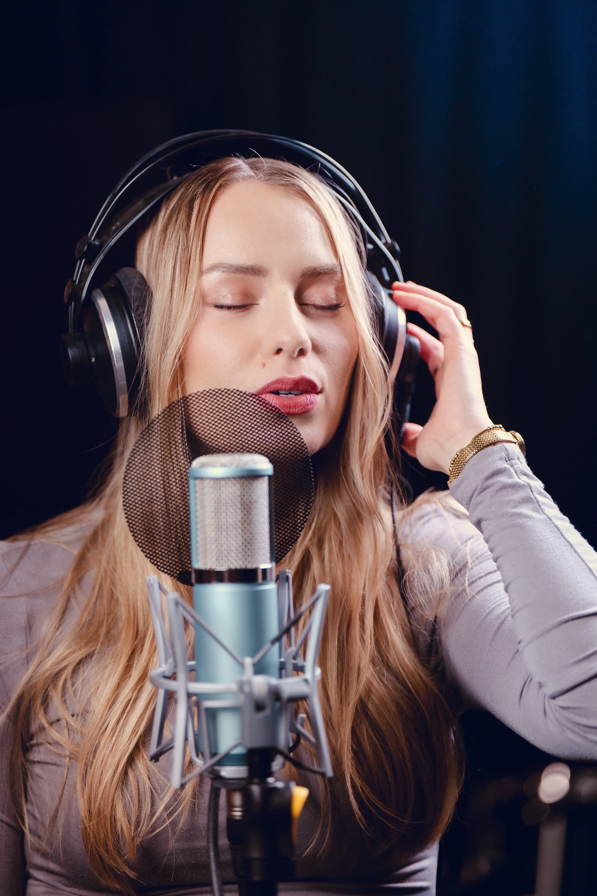 A woman with long blonde hair wearing headphones, singing into a professional microphone in a recording studio.