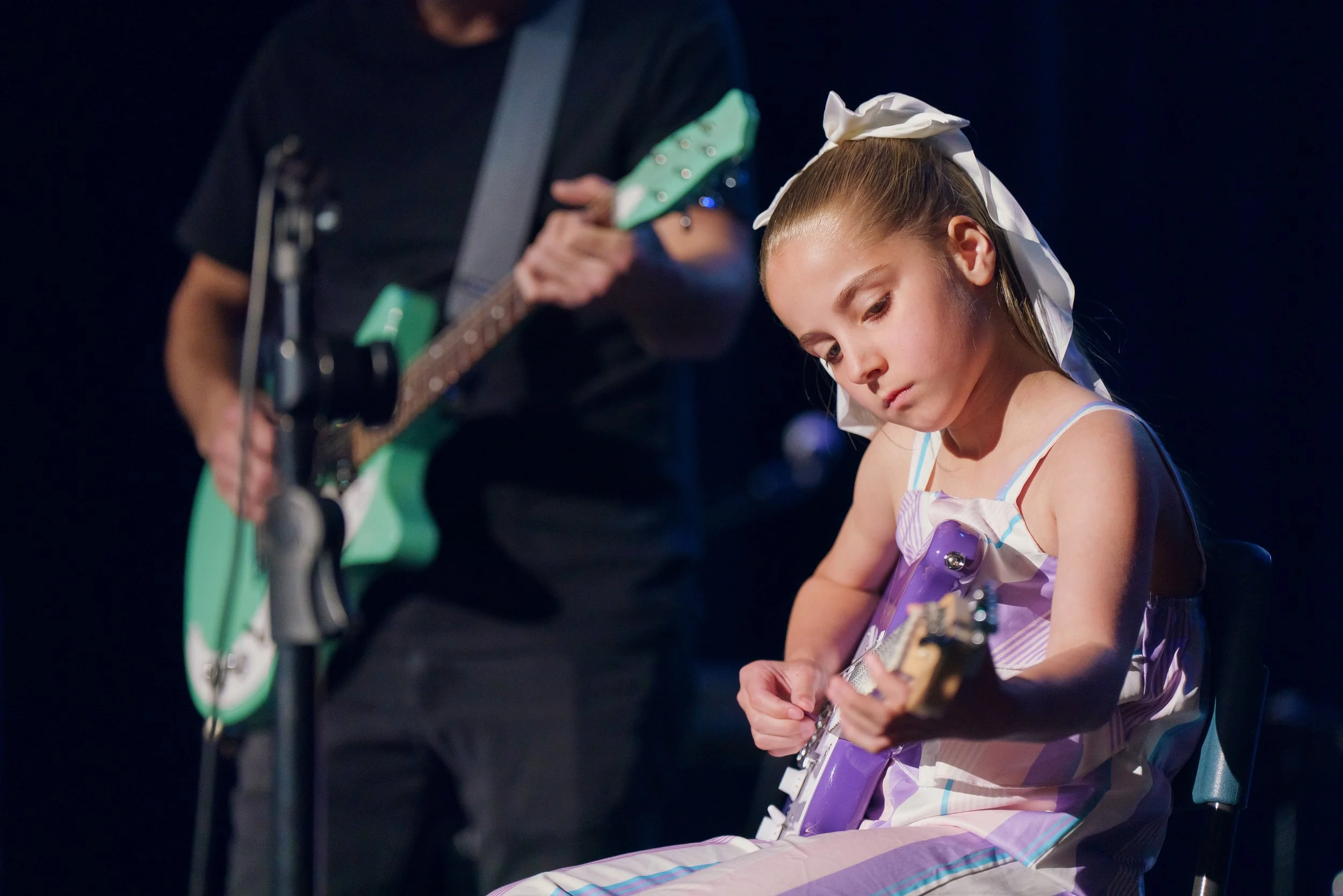 A young girl playing an electric guitar on stage with a person in the background playing an acoustic guitar.
