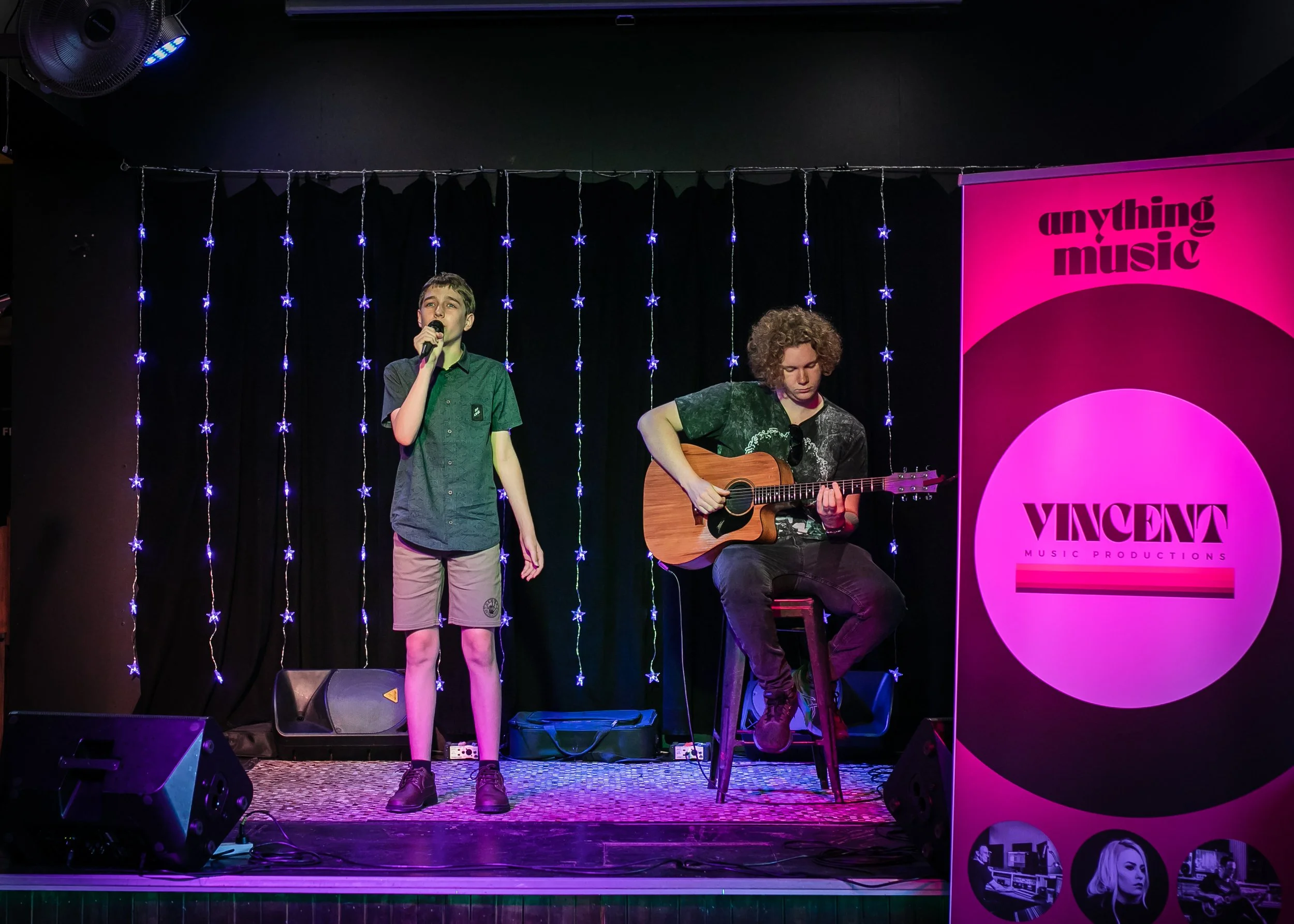 Two young male performers on a stage, one singing into a microphone and the other playing an acoustic guitar. The stage is decorated with string lights and there is a pink banner that reads 'Vincent Music Productions' with the slogan 'anything music'