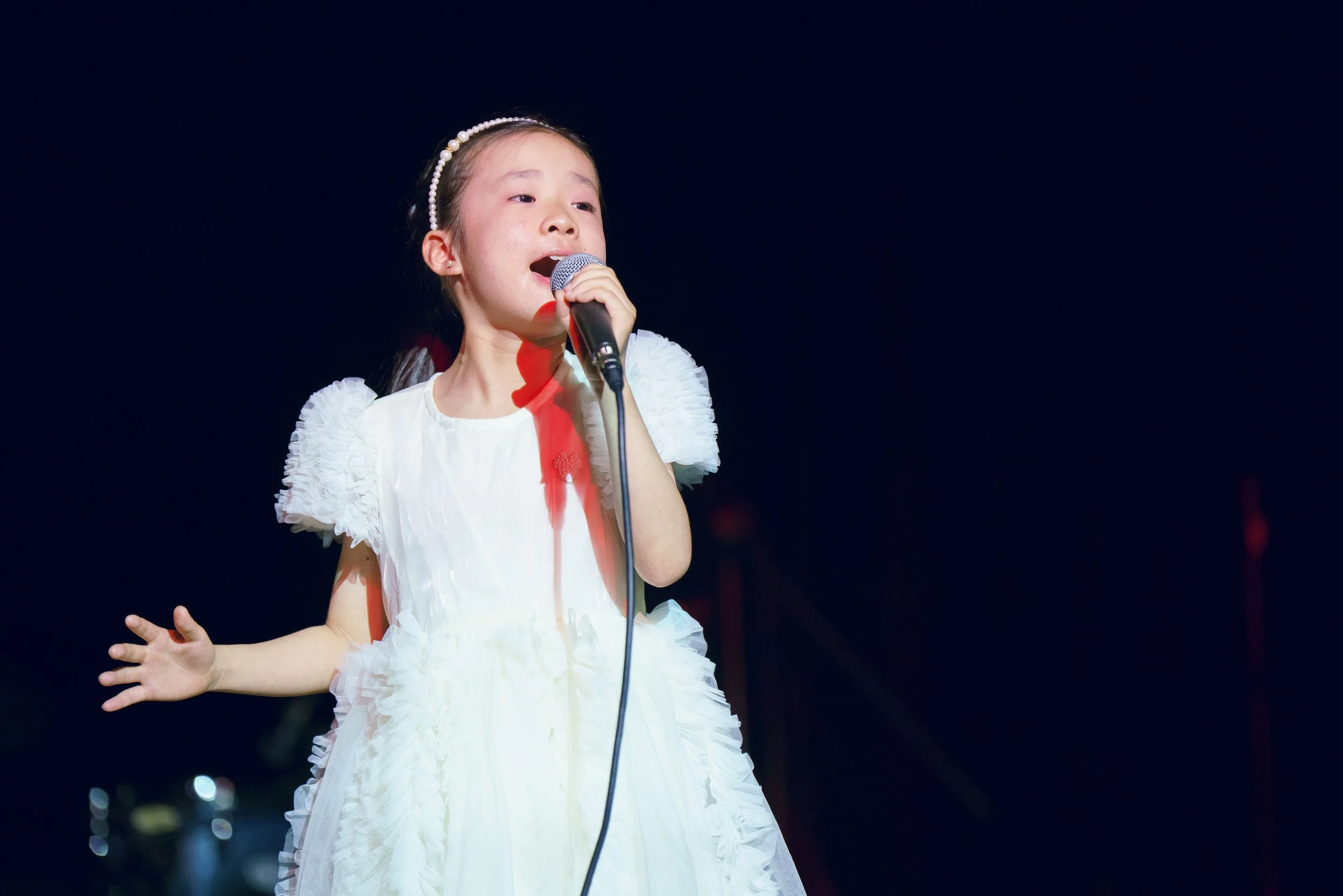A young girl in a white dress singing into a microphone on stage.