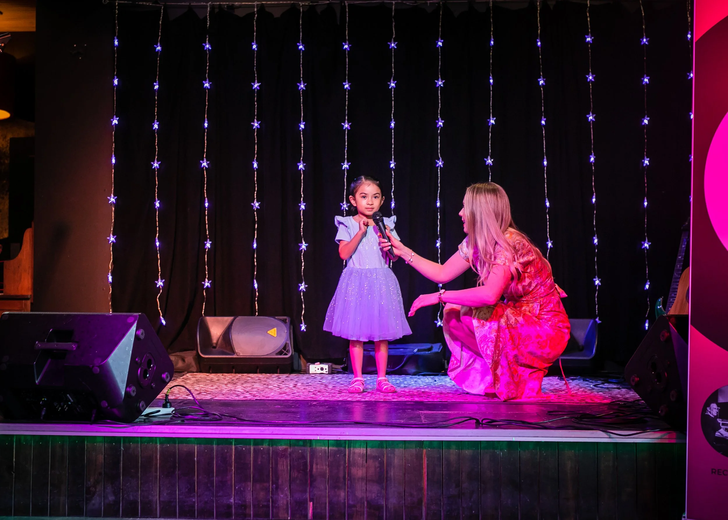 A young girl in a purple dress standing on a stage, holding a microphone, as a woman in a red dress kneels next to her during a performance with decorative string lights in the background.