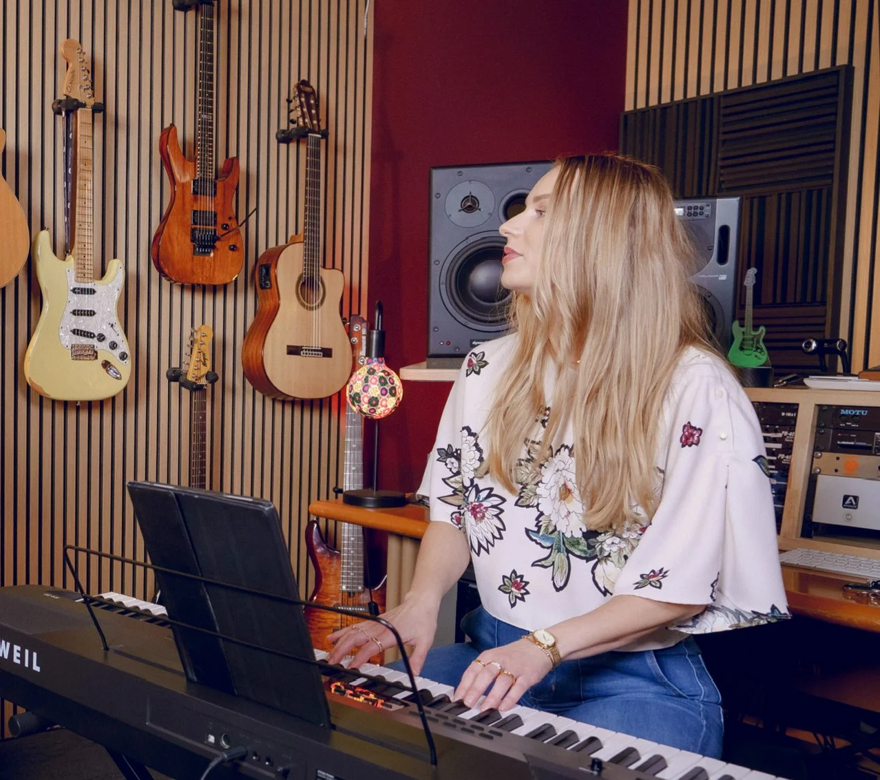 A woman playing a digital keyboard in a music studio with guitars hanging on a wooden wall behind her.
