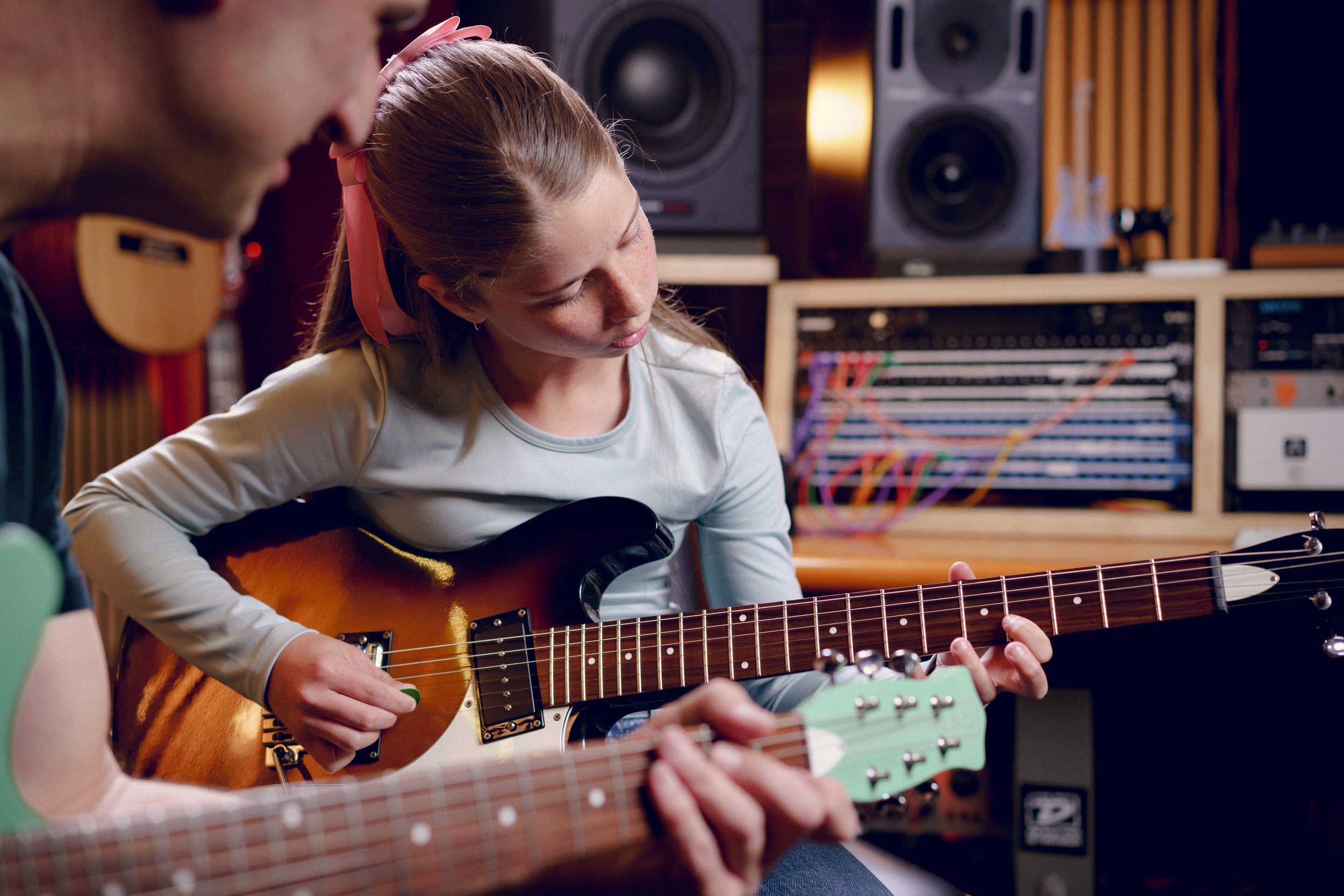 A young girl and a man playing electric guitar together in a music studio, with audio equipment and sound mixing gear in the background.