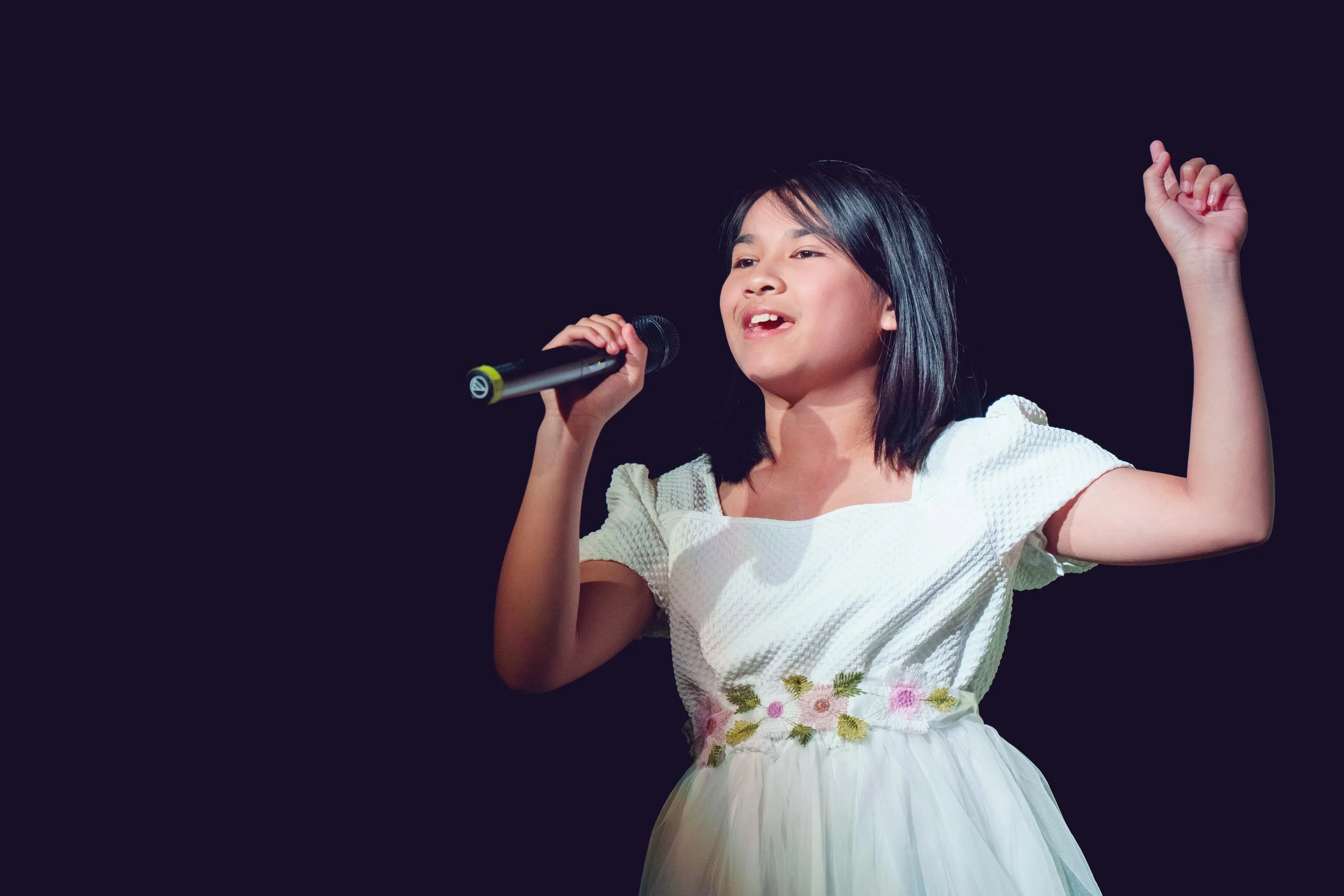 Young girl singing into a microphone with one hand raised, wearing a white dress with floral embroidery, against a dark background.