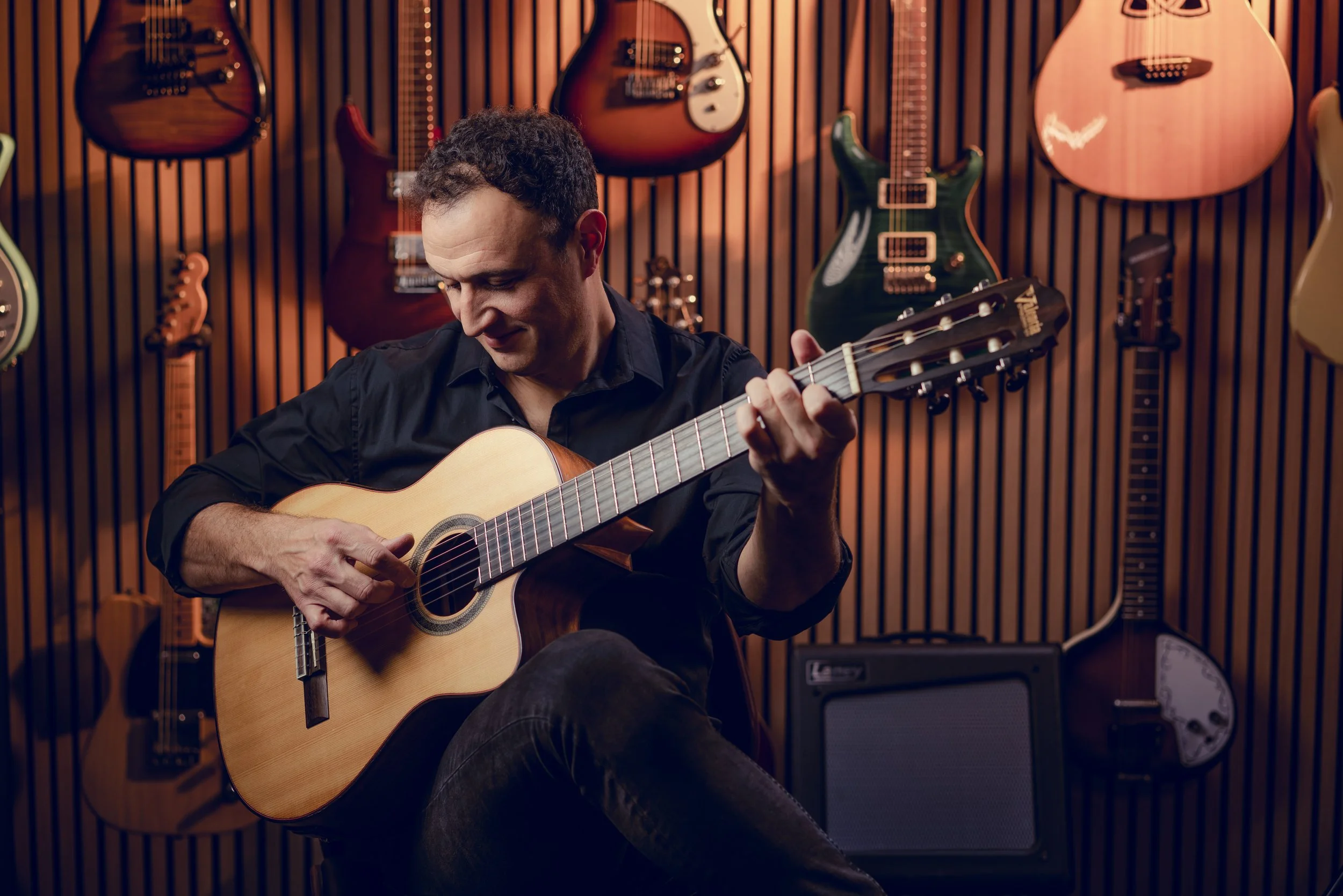 A man playing an acoustic guitar in a music store with multiple guitars hanging on the wall behind him.