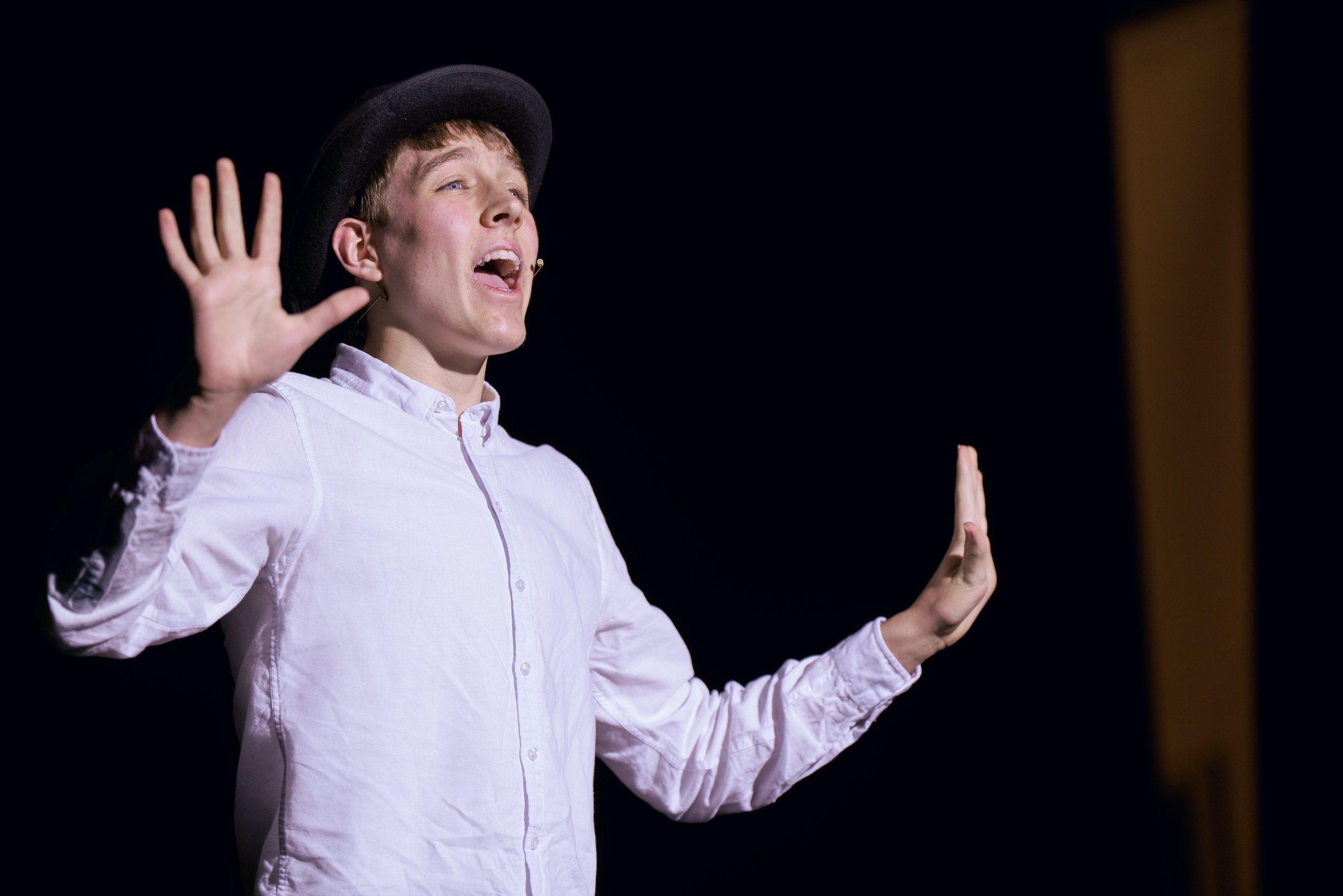 A young man wearing a black hat and a light pink button-up shirt is speaking or singing on stage with his eyes closed, raising both his hands with palms facing outward against a black background.