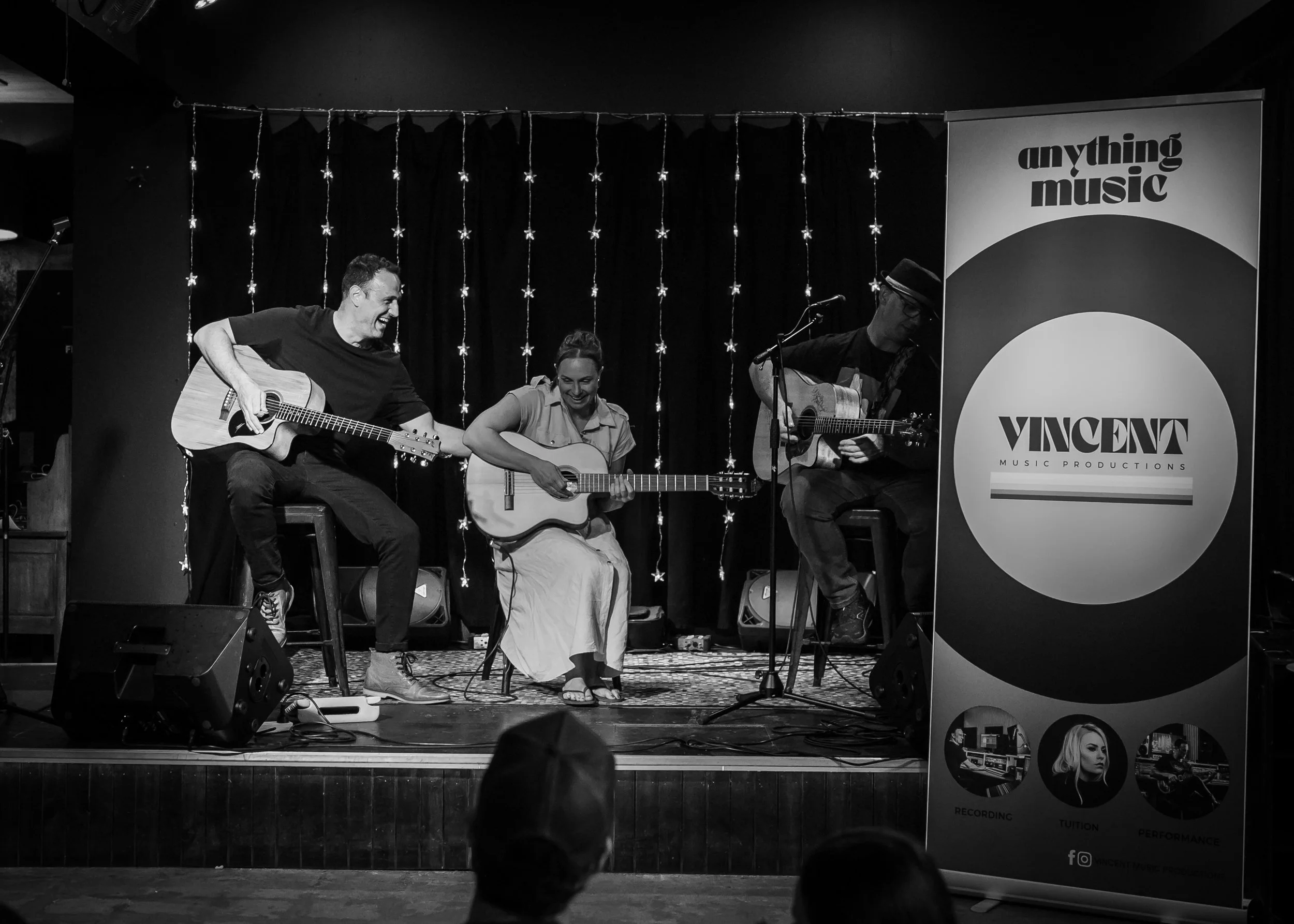 Three musicians perform on a small stage with guitars, smiling and playing music. A company banner with the text 'VINCENT MUSIC PRODUCTIONS' stands to the right. The background has string lights and a black curtain.