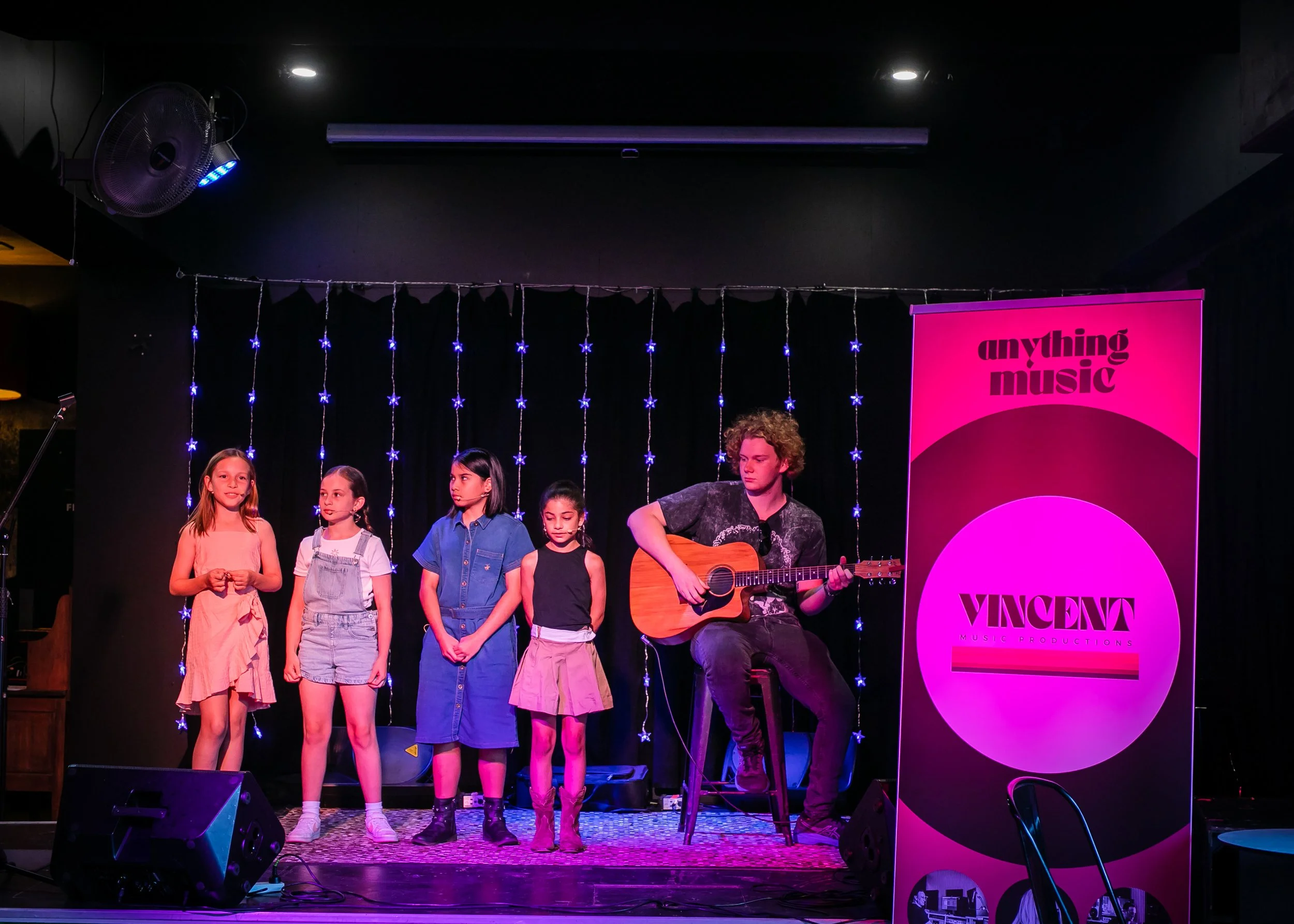A group of children and a young man with curly hair playing guitar on stage, with a pink and purple banner reading 'anything music' and 'Vincenza music productions' behind them, and string lights hanging in the background.