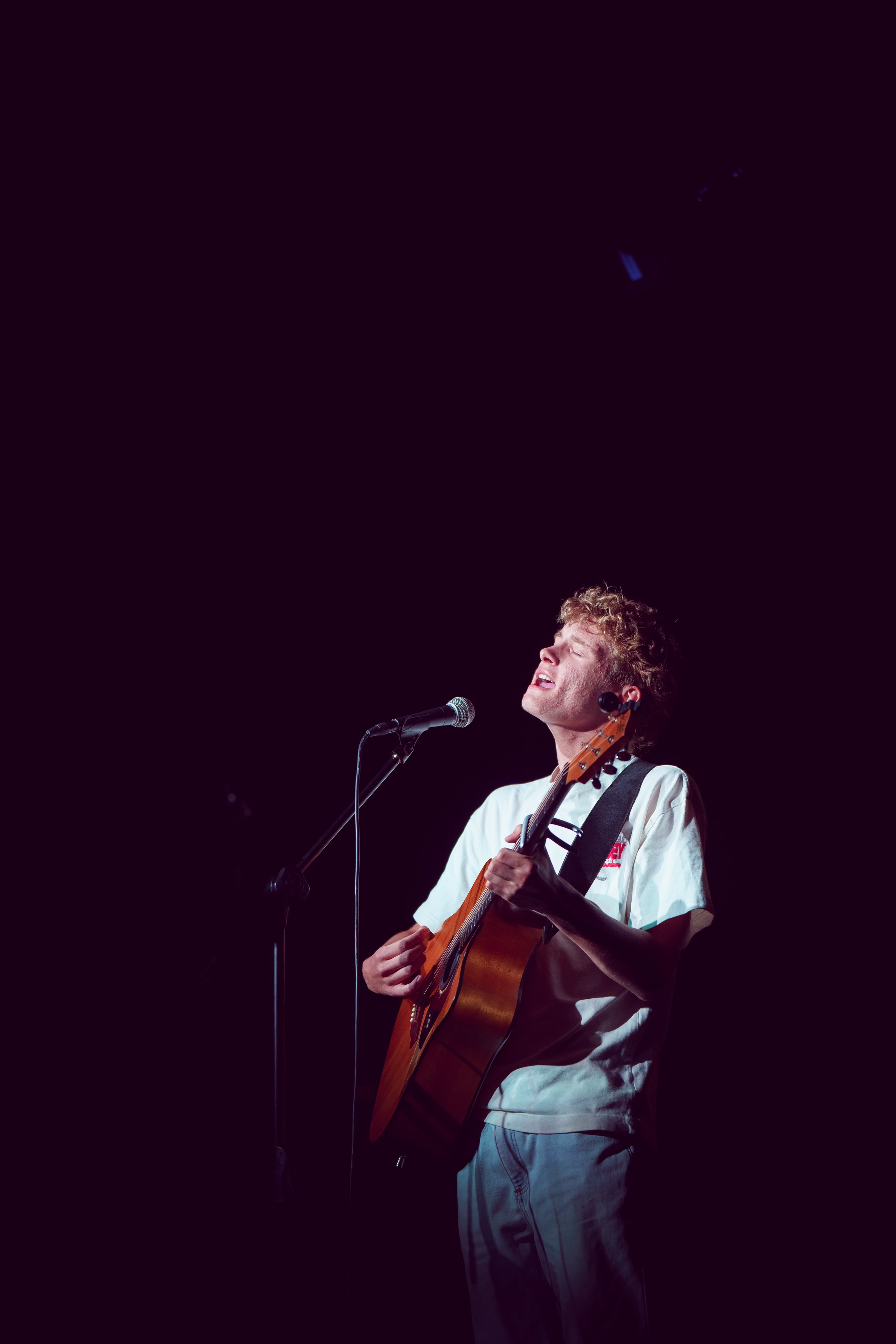 Young man performing passionately with his eyes closed, playing an acoustic guitar and singing into a microphone on a dark stage.