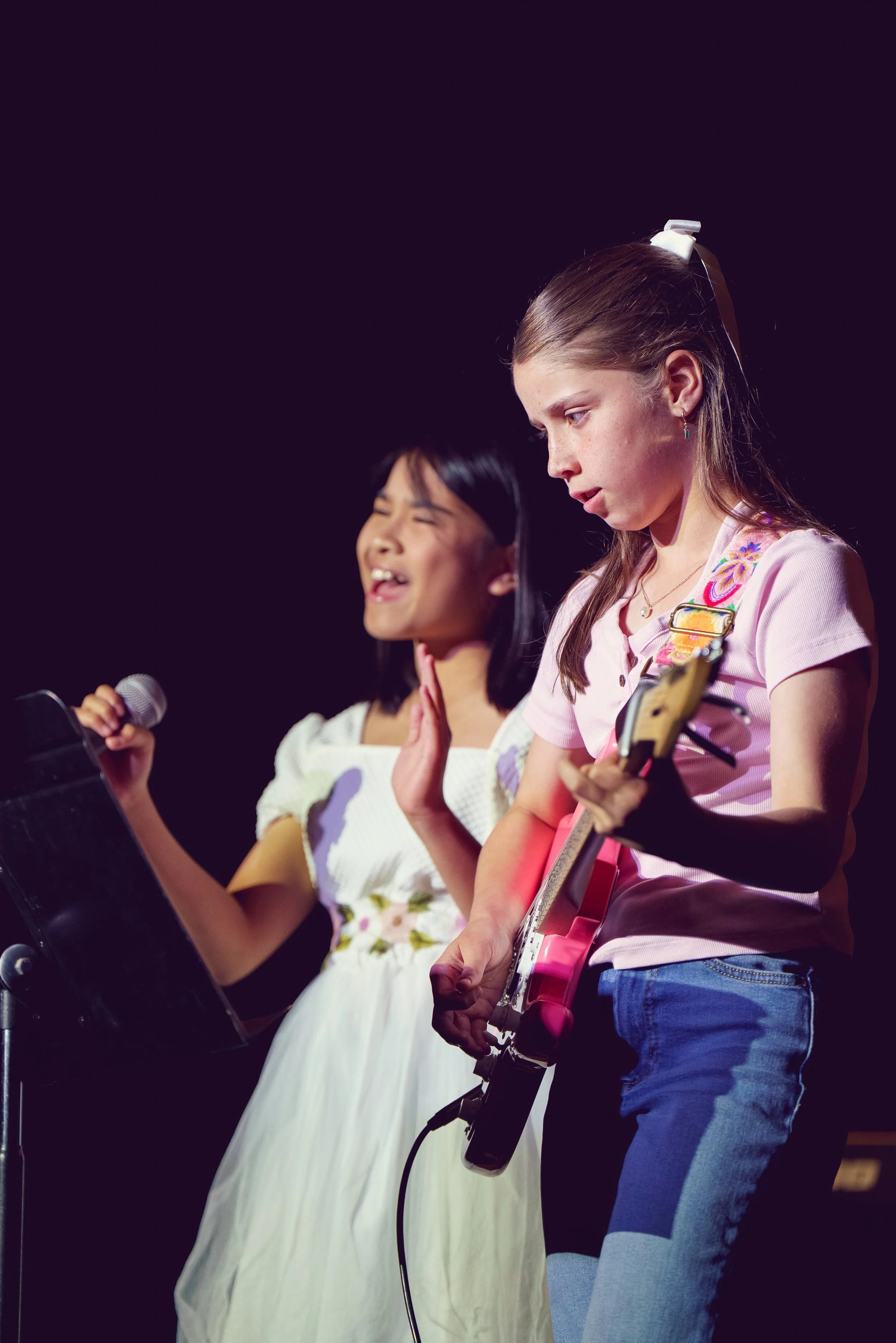 Two young girls performing on stage, one singing into a microphone and the other playing an electric guitar.