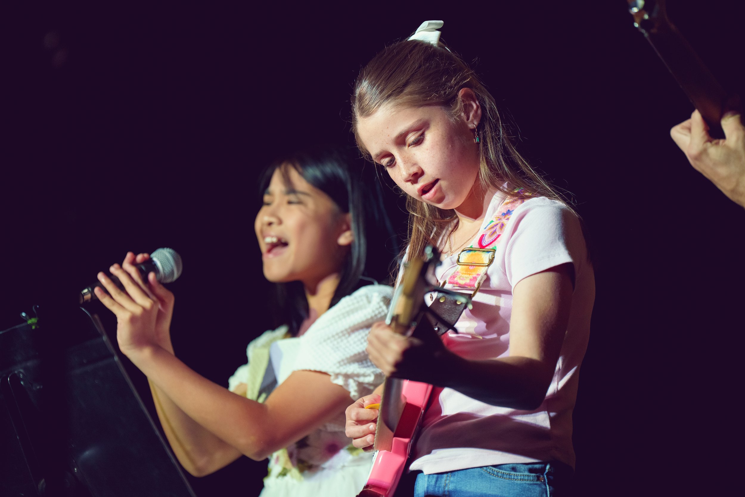 Two young girls, one singing into a microphone and the other playing a guitar, performing on a stage with a dark background.