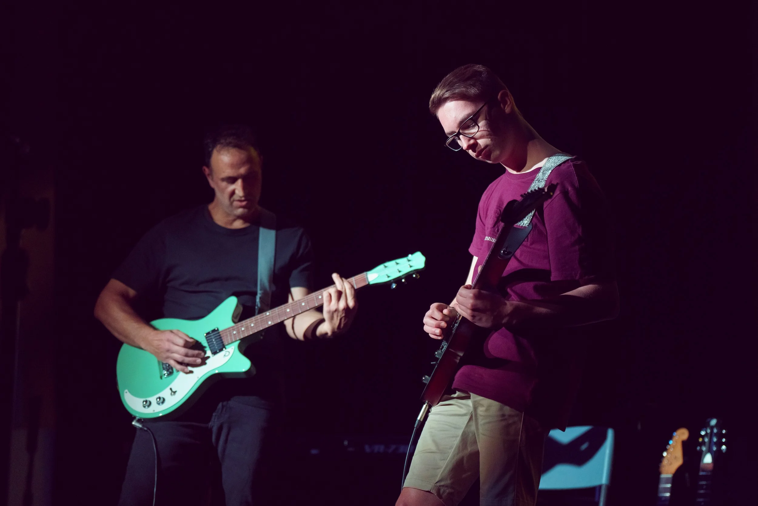 Two musicians playing electric guitars on stage during a performance, with dark background.