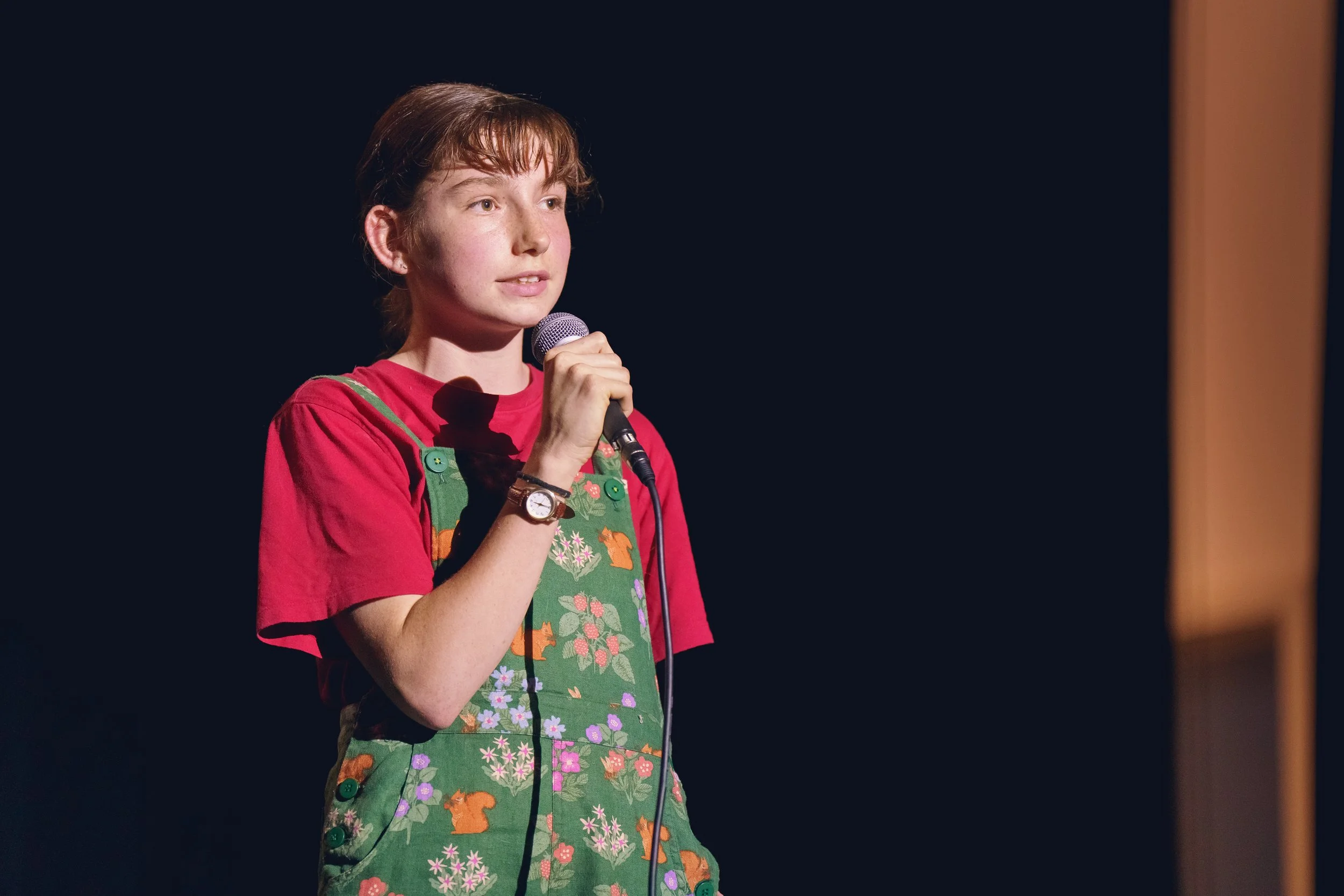 A young girl with red hair wearing a red shirt and green floral overalls holding a microphone on stage with a black background.