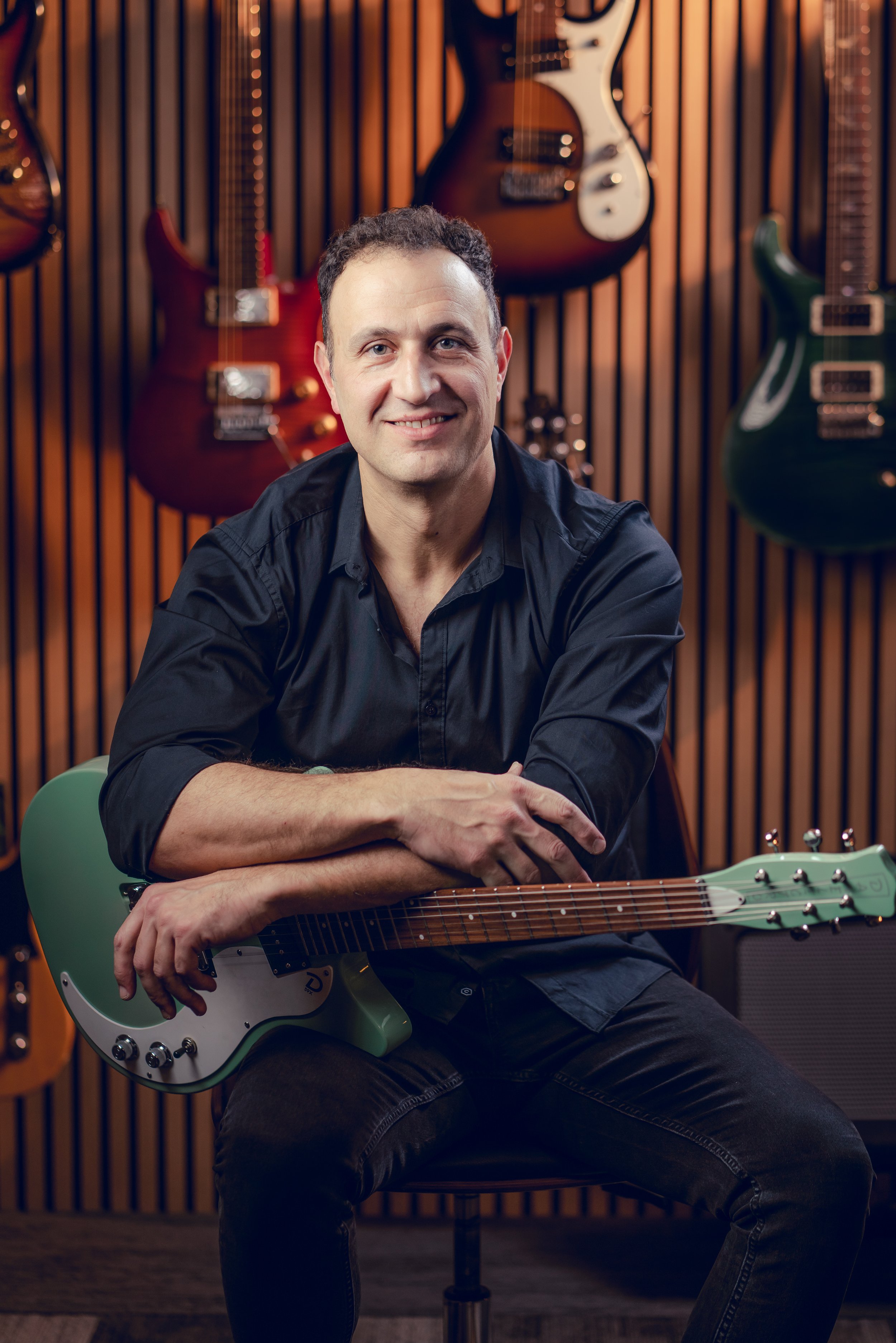 A man with short dark hair smiling at the camera, sitting with a green electric guitar across his lap in a music studio with various guitars hanging on a wooden wall behind him.