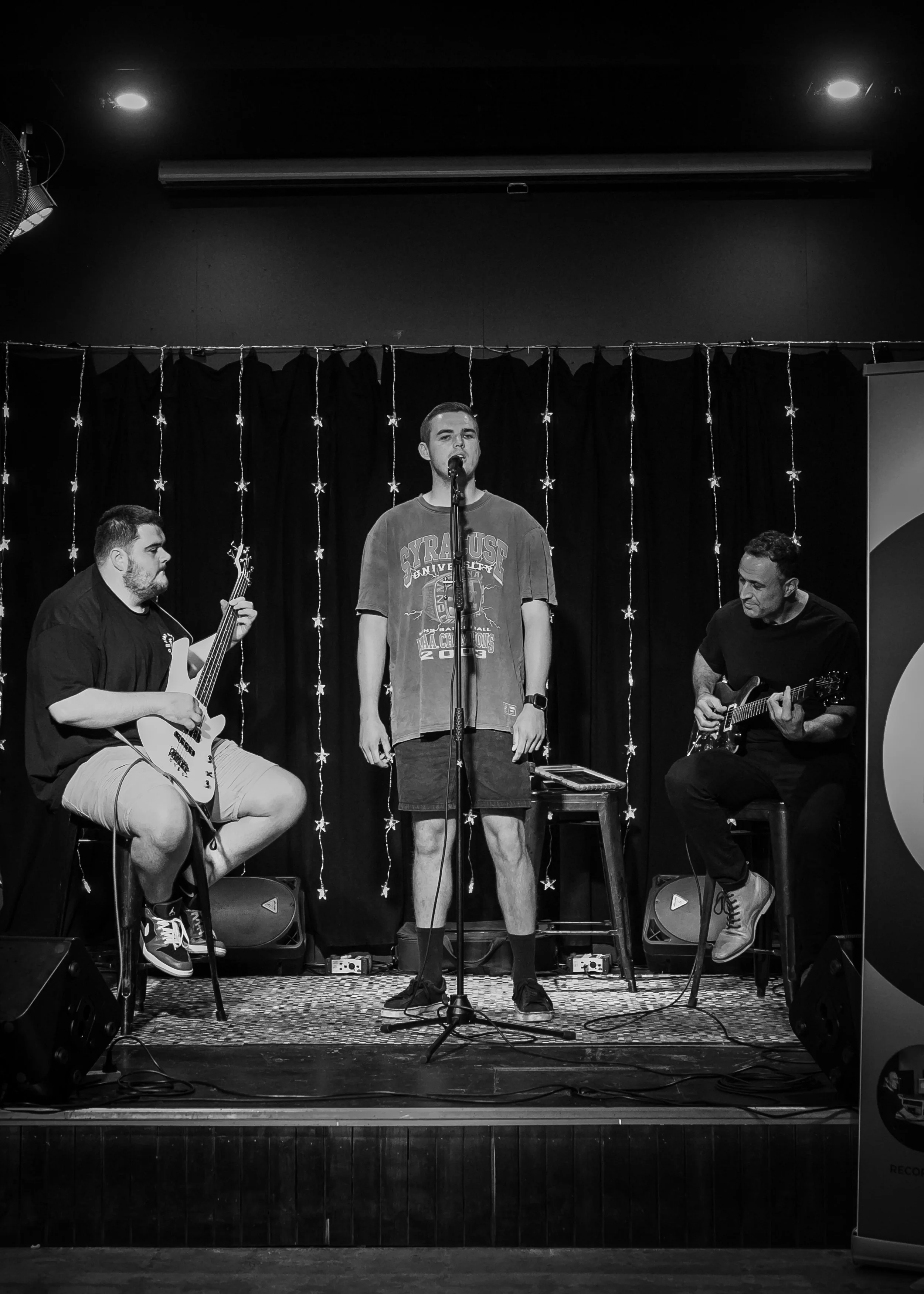 Three men performing music on a stage with black curtains and star-shaped string lights. Two are playing guitars, and one is singing into a microphone.