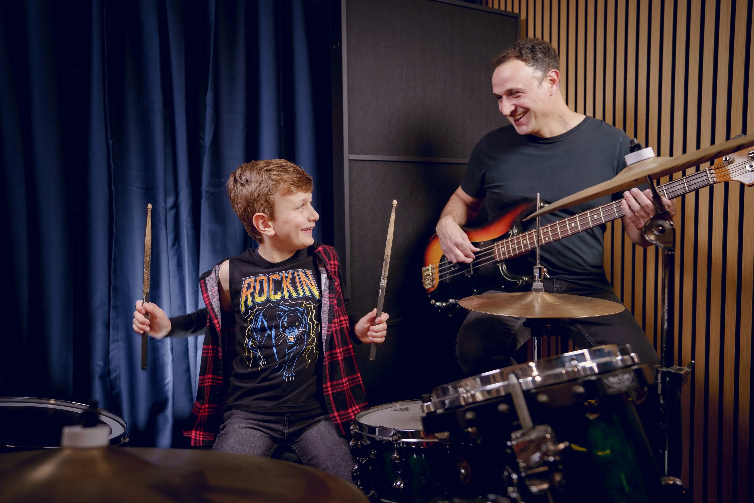 A young boy and an adult male musician share a moment in a music rehearsal. The boy, wearing a black t-shirt with a rock-themed design and plaid shirt, holds drumsticks and smiles at the man. The man, sitting on a stool, holds a bass guitar, smiling back. They are in a room with dark walls and blue curtains.