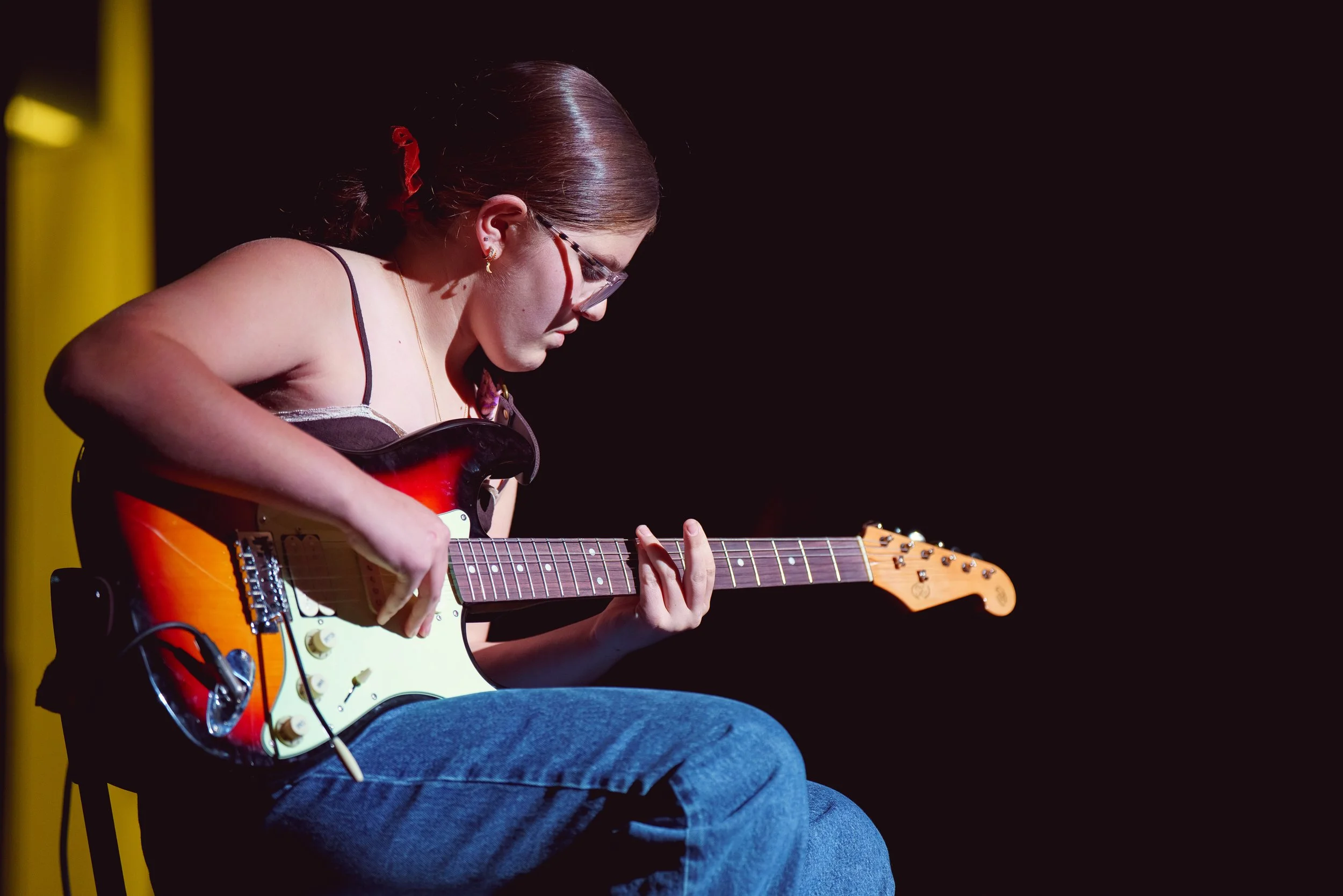 Young woman with glasses playing an electric guitar on a dark stage.